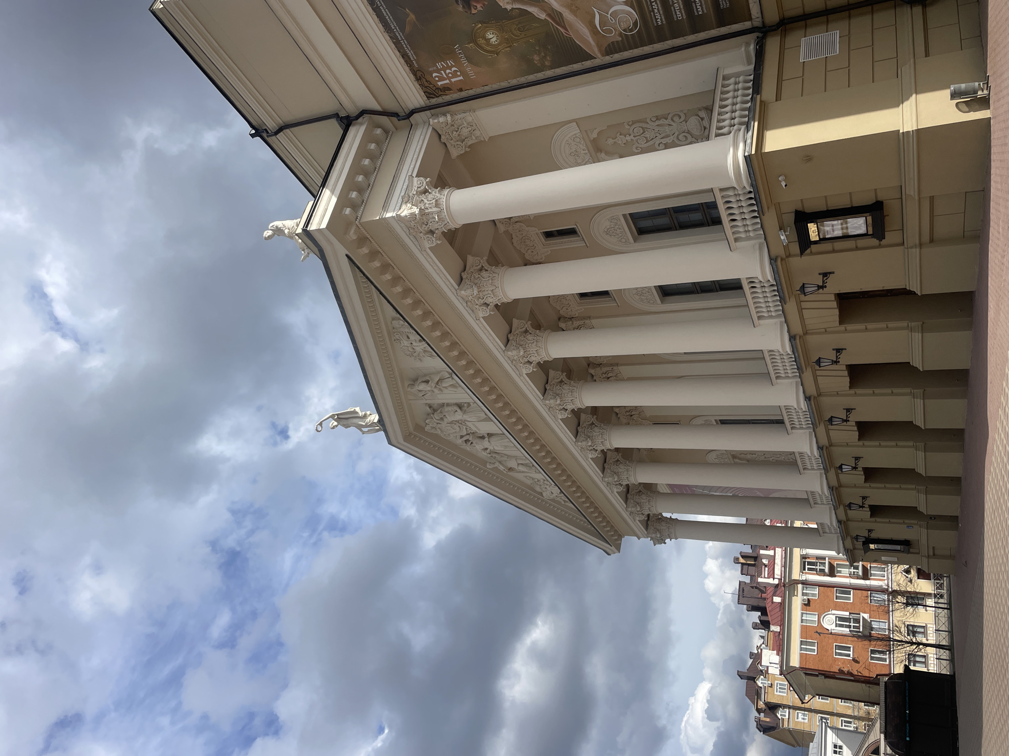 The image, rotated 90 degrees clockwise, captures a grand, classical-style building from a low-angle perspective, looking up towards its ornate facade and the sky. The building, likely a theater or public institution in Kazan, Russia, features a prominent portico with a series of tall, white, fluted columns topped with elaborate Corinthian capitals. Above the columns, an intricately carved frieze depicting figures and decorative motifs runs along the facade. Stylized white figures, resembling decorative sculptures or gargoyles, are positioned on the corners of the pediment, silhouetted against the dramatic sky.
The building's exterior is a light cream or beige color, complemented by darker architectural accents. Large promotional banners or posters are affixed to the upper part of the building, above the columned portico. Visible Cyrillic text on these posters includes "13 MApTA" (March 13), "ПРЕМЬЕРА" (Premiere), "НАДЕЖДА" (Nadezhda / Hope), and "СЕРГЕЙ" (Sergey). A prominent "25" is also visible, possibly indicating an anniversary, along with a classical portrait of a woman.
Below the main portico, the building facade shows windows, and sconce-style lights are attached to the walls, along with a small display case. The sky is overcast with large, dramatic grey and white clouds, suggesting a partly cloudy or impending stormy weather condition. To the lower right, beyond the main building, other urban structures with varying architectural styles, including multi-story buildings with red brick and lighter facades, are visible. A paved area is seen in the foreground on the right. The scene depicts daytime, with natural light illuminating the architecture. No specific human activity is shown.
