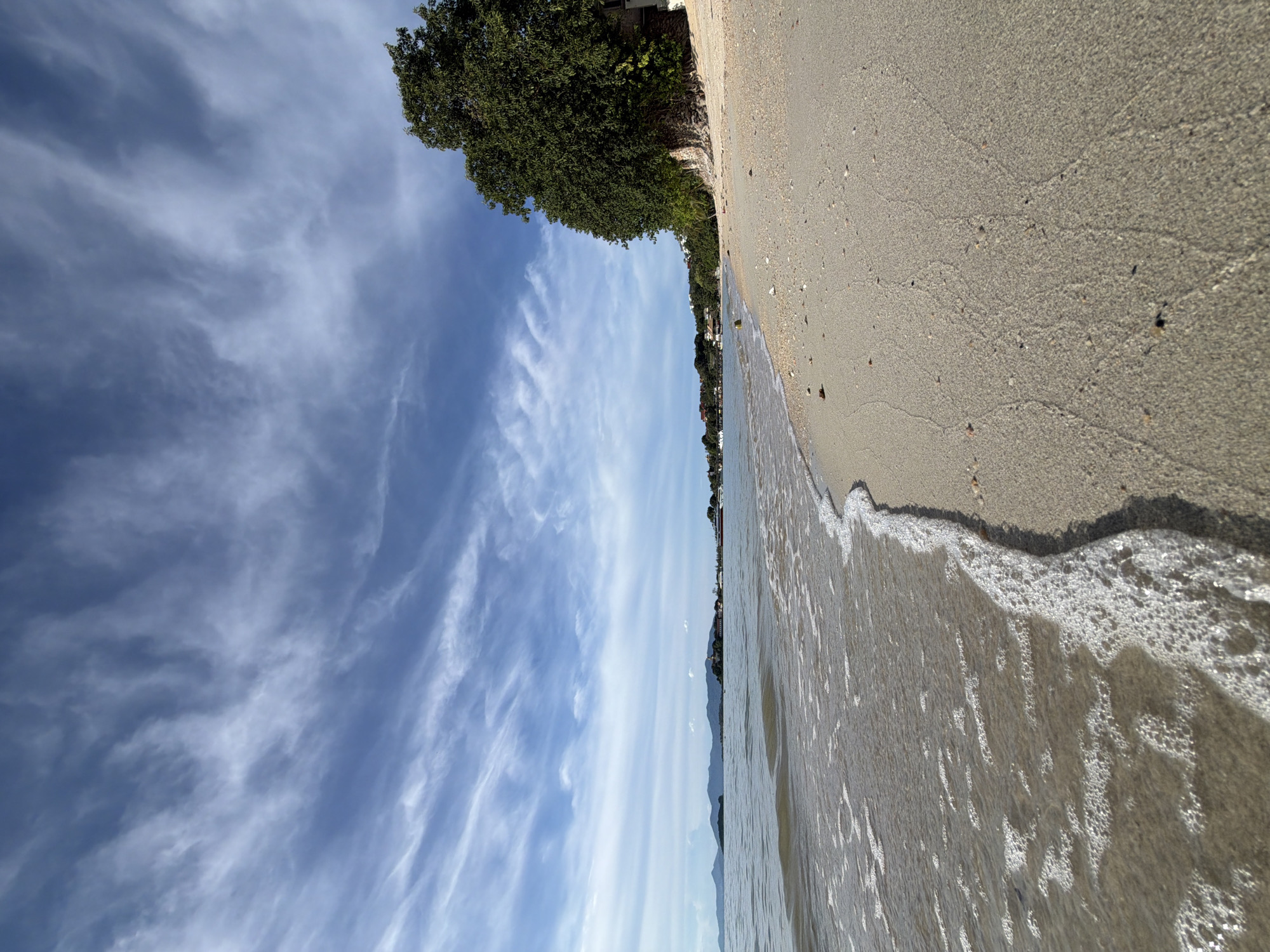 The image depicts a tranquil beach scene in Baan Bo Phut, Thailand. The foreground is dominated by the sandy shore, with gentle waves lapping at the edge, creating a foamy white line along the waterline. The sand itself is light brown and appears relatively smooth, marked by subtle ripples from the receding water.
To the left of the frame, a vibrant green tree stands tall, its dense foliage contrasting with the soft hues of the sky and sand. Beyond the tree, a line of buildings and vegetation stretches along the coastline, suggesting a developed area adjacent to the natural beach. In the distance, faint mountain outlines can be seen against the horizon.
The sky is a brilliant blue, adorned with wispy white clouds that stretch across the expanse. The lighting suggests it is daytime, likely late morning or early afternoon, with the sun casting a soft glow over the scene. The overall atmosphere is peaceful and serene, conveying a sense of calm and natural beauty. There is no visible text in the image.