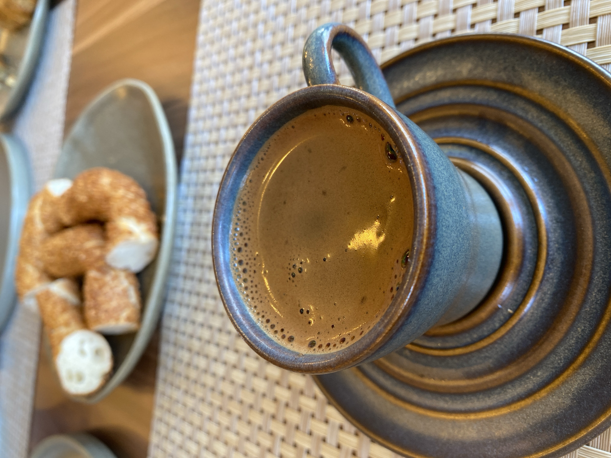 In Sokhumi, Abkhazia, a close-up shot captures a cup of coffee and a pastry on a table. The coffee, a rich brown with a frothy top, sits in a rustic blue ceramic cup. This cup rests on a matching saucer with concentric rings. In the background, a light-colored plate holds slices of a ring-shaped pastry, possibly a simit, dusted with sesame seeds. The setting appears to be a cafe or restaurant, with a woven placemat and a wooden table visible. The lighting is natural, highlighting the textures of the food and ceramic ware. There are no people visible in the frame.