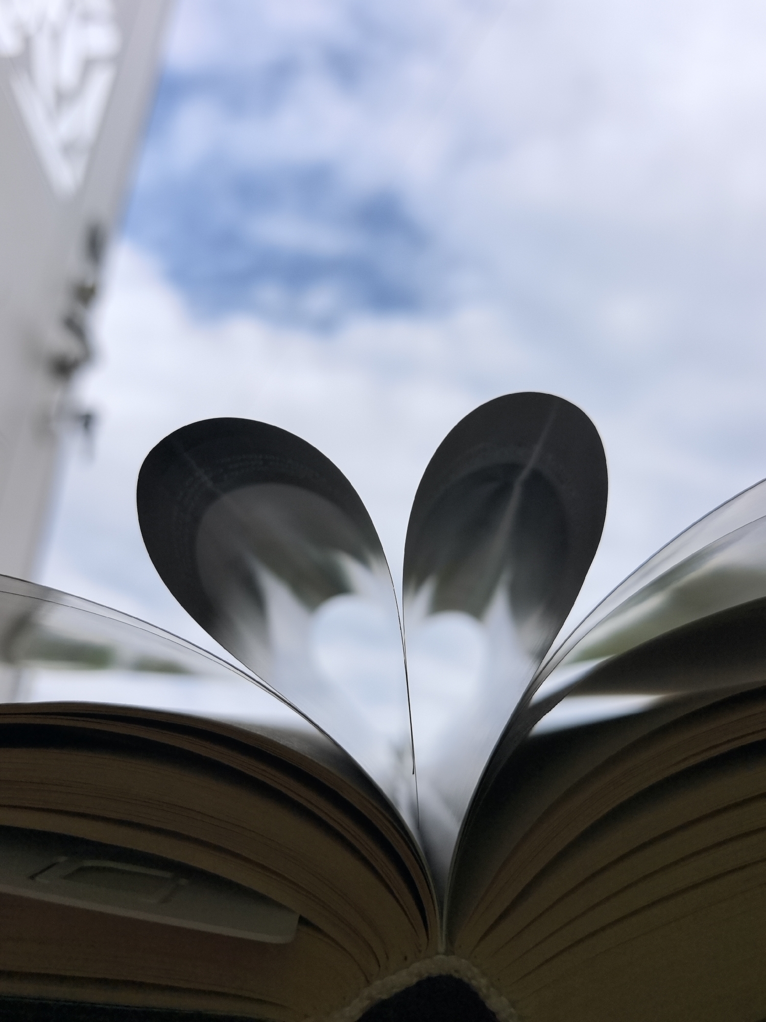 The image shows an open book with pages folded to create a heart shape. The background is a blurred view of a cloudy sky and what appears to be part of a building, possibly a balcony railing. The book has aged pages, and there's a bookmark visible among them. The focus is on the heart shape formed by the pages and the sky visible through it. The city of Noviy, Russia, is not identifiable within the image.