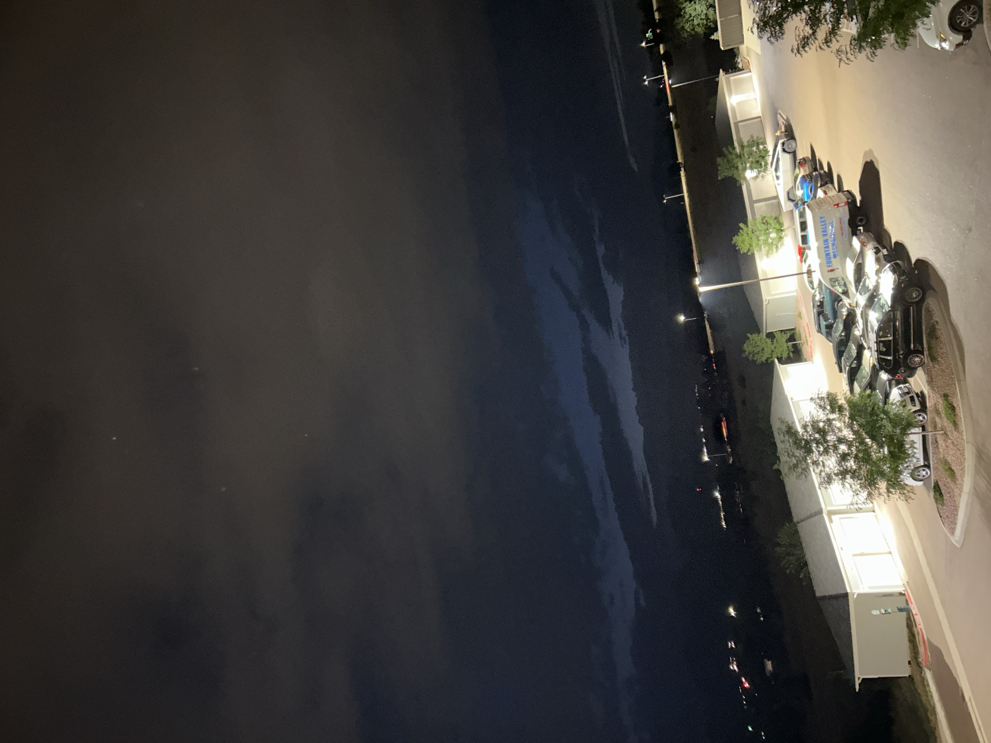 An exterior, high-angle, night-time photograph captures a dimly lit parking lot adjacent to a low-rise, multi-unit residential building in Colorado Springs, USA. The sky is dark and filled with heavy clouds, with a few distant lights from the city visible on a dark, undulating horizon. In the foreground, a row of parked vehicles is illuminated by overhead streetlights, including a white van with lettering that reads "FOUNTAIN VALLEY MECHANICS" and a silver Tesla Cybertruck. Several trees are scattered throughout the parking lot and along the edge of the property, some of which are bare, suggesting a deciduous tree in an autumn or winter season. The building appears to be a garage or storage facility, with several large bay doors visible. The overall atmosphere is one of a quiet, residential area at night.