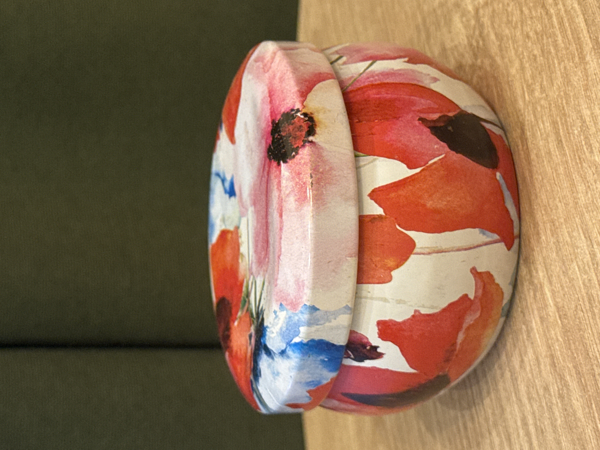 Two cylindrical, flower-patterned tins sit on a light brown surface. The tins are oriented vertically. The tin on the left displays red, white, and blue floral designs, while the tin on the right features predominantly red floral patterns on a white background. A dark green surface is visible to the left of the tins. The location is Unknown, Unknown.