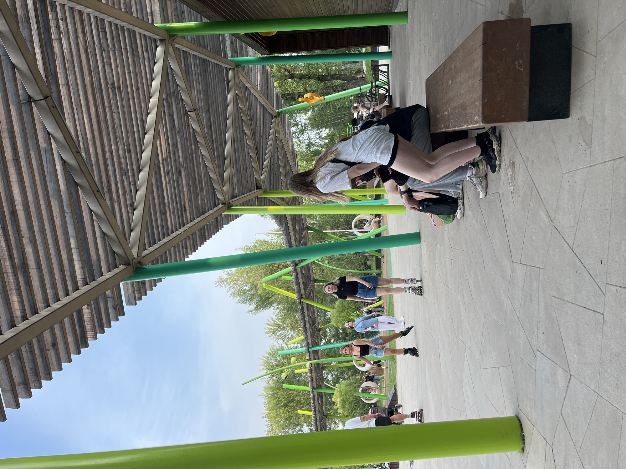 The image depicts an outdoor recreational area with a modern, open-air shelter featuring a wooden slatted roof supported by vibrant green cylindrical pillars. The ground is paved with large, irregularly shaped concrete tiles. In the foreground, a wooden bench is occupied by several individuals, their legs and feet visible. A young woman in a white t-shirt and black shorts is standing, facing away from the camera, with her right leg bent at the knee, suggesting movement or a pose. She is wearing black and white sneakers. Behind her, other individuals are seated on the bench, partially obscured.
Further into the scene, several people are engaged in rollerblading and skateboarding. Two women are rollerblading, one in a black tank top and denim shorts, the other in a black t-shirt and denim shorts. A person on a skateboard is visible on the left. In the background, there are trees and a playground-like structure with bright green and yellow elements. The sky is partly cloudy.
This location is identified as the "Stolby National Park" (Национальный парк "Красноярские Столбы"), specifically in the area near the "Funicular Entrance" (Вход на фуникулер) in Krasnoyarsk, Russia. This park is known for its unique rock formations and recreational facilities. The architecture of the shelter and the general ambiance suggest a developed tourist or recreational zone within the park.