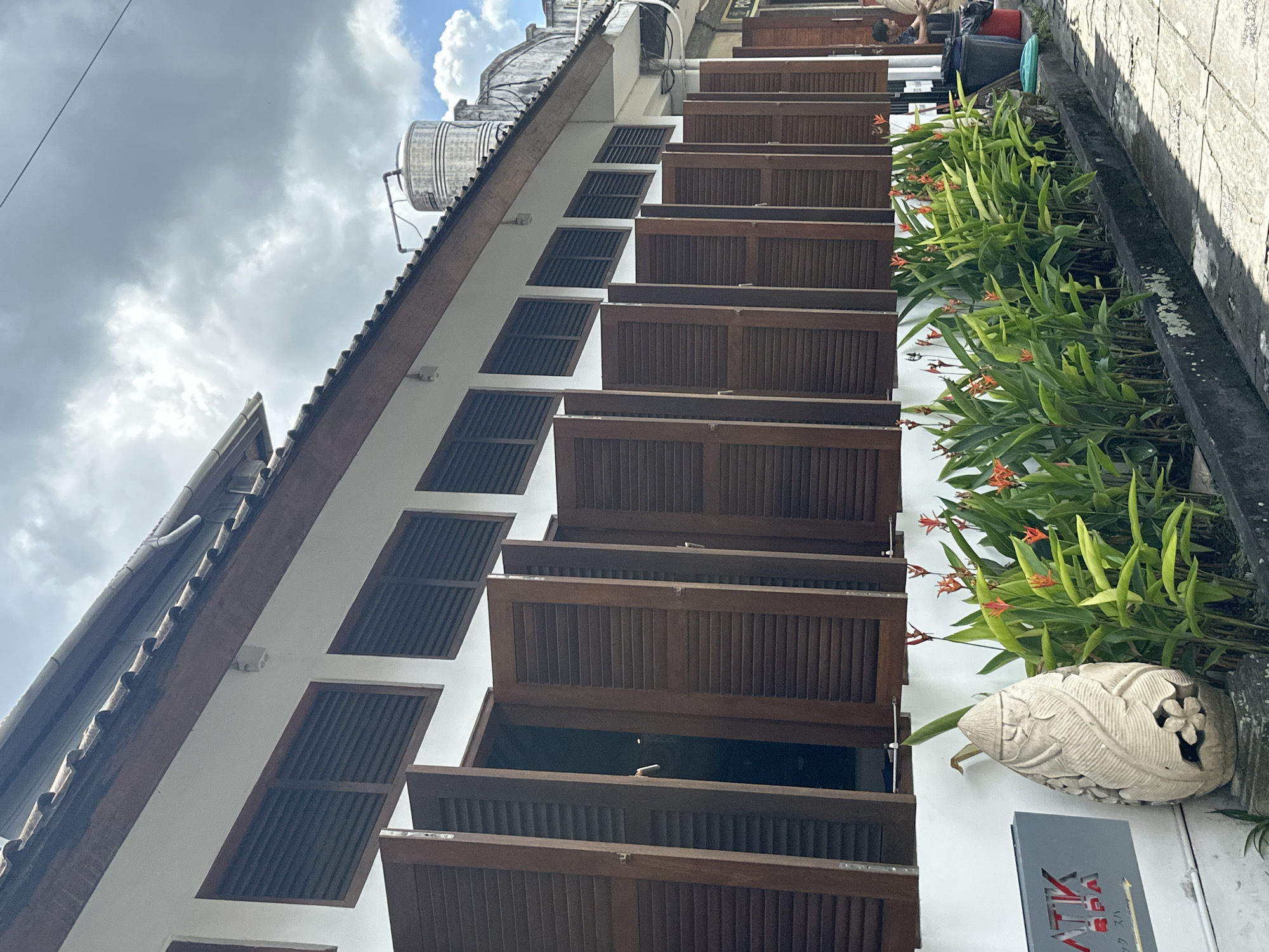 In Ubud District, Indonesia, a row of wooden louvered shutters are open along the side of a white building with a brown tiled roof. Lush green plants with orange flowers are planted in a bed along the exterior wall, and a carved stone ornament stands next to a sign that reads "ATIK SPA". Further down the street, a person is visible near a water tank on the roof of another building. The sky is cloudy with patches of blue.