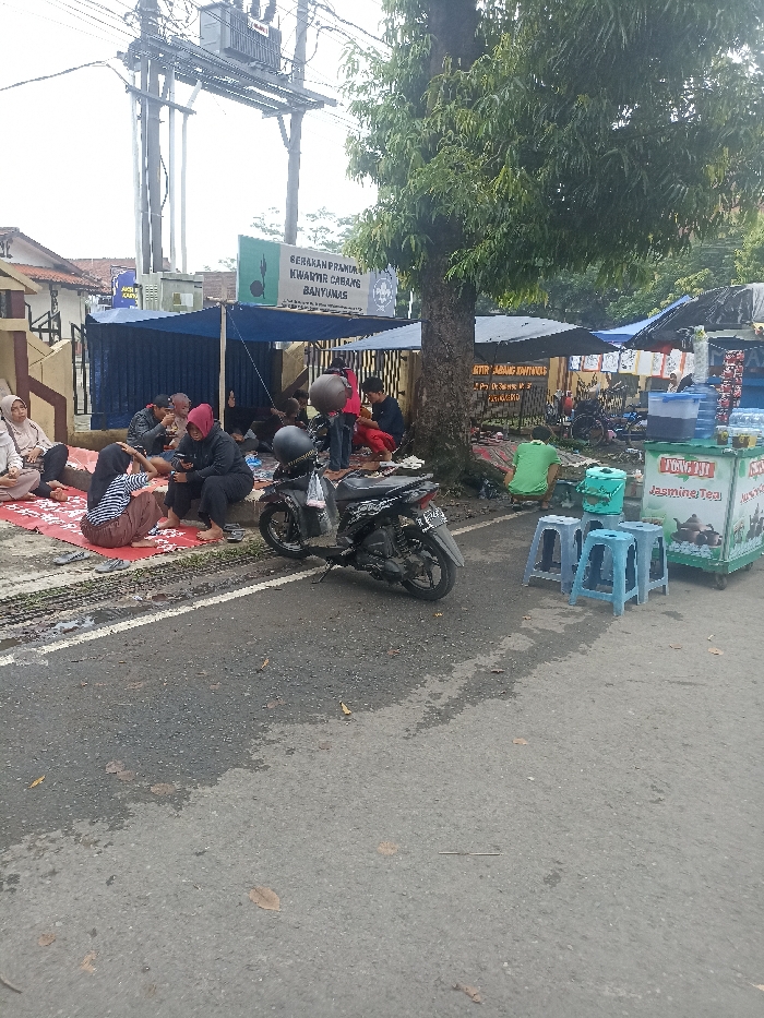 The scene shows a group of people sitting on the ground in front of a street food vendor, located near a large tree at the corner of a street, on the street of Jalan Jenderal Soedirman, Purwokerto, Indonesia. They are all dressed in casual clothing. There is a small motorbike parked nearby and a sign in Indonesian that reads "Serakan Pramuka Kwartir Cabang Banyumas." The food vendor is selling a variety of drinks, including "Tofu Pai Jasmine Tea." The ground is paved with asphalt, and the surrounding area is a mix of concrete buildings and trees. The overcast sky suggests the day is cloudy.