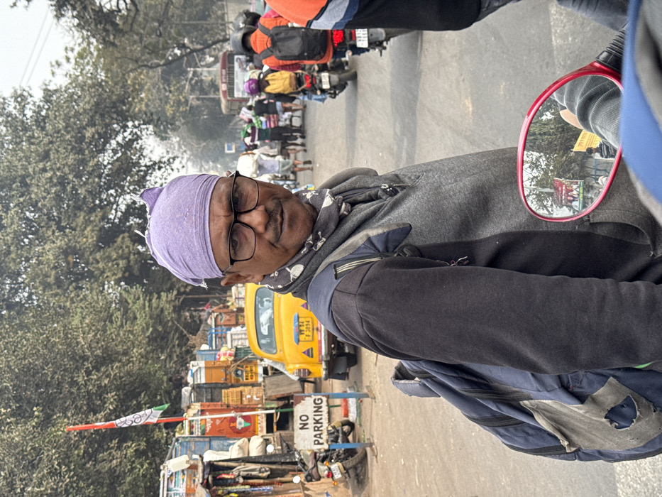 An individual wearing a purple head covering and glasses is seated on a vehicle, possibly a scooter, with a red side mirror visible. The background shows a busy street scene with trees, a yellow taxi, and various vehicles. A "NO PARKING" sign is prominent. The image seems to be taken from the individual's perspective while on the move.