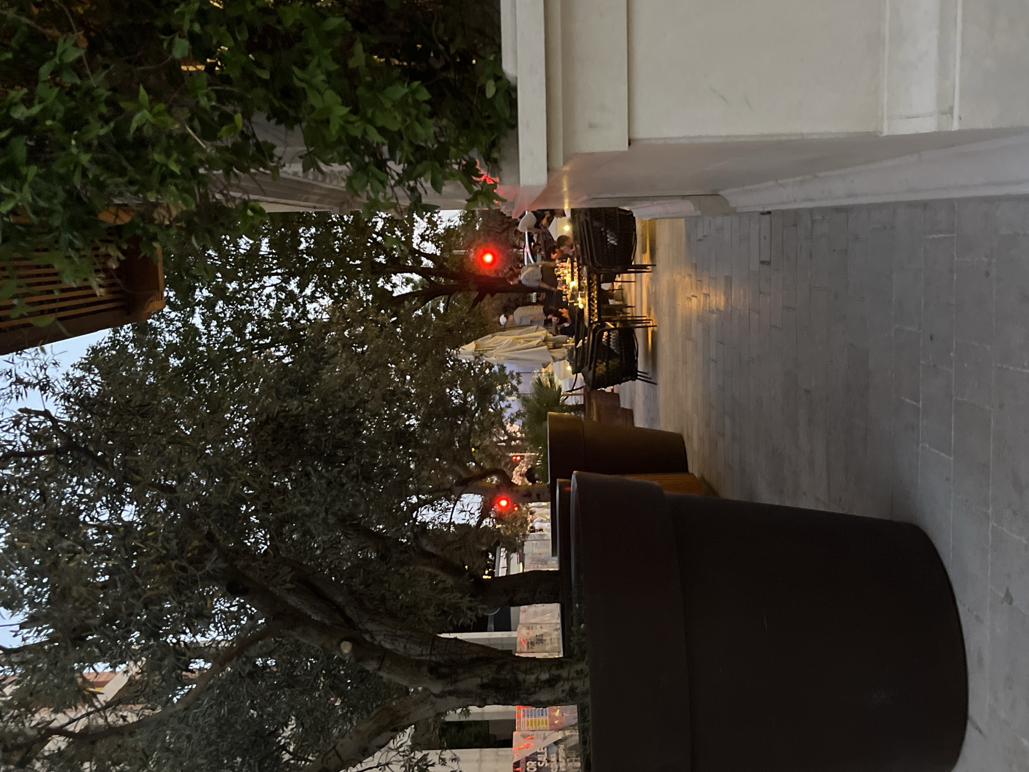 The image depicts an outdoor seating area of a restaurant or cafe in Budva, Montenegro, likely during early evening hours. A large, dark brown planter pot dominates the foreground on the left, obscuring a significant portion of the view. To the right of the planter, a light-colored stone wall is visible, with lush green foliage growing on top.
Beyond these elements, a paved walkway leads to a seating area where several tables and chairs are arranged. People are seated at these tables, engaged in conversation and dining. The tables are illuminated by warm, ambient lighting, creating a cozy atmosphere. In the background, trees with dark foliage are scattered, and a red traffic light is visible, suggesting the proximity of a road. The overall scene suggests a relaxed dining experience in an urban setting.