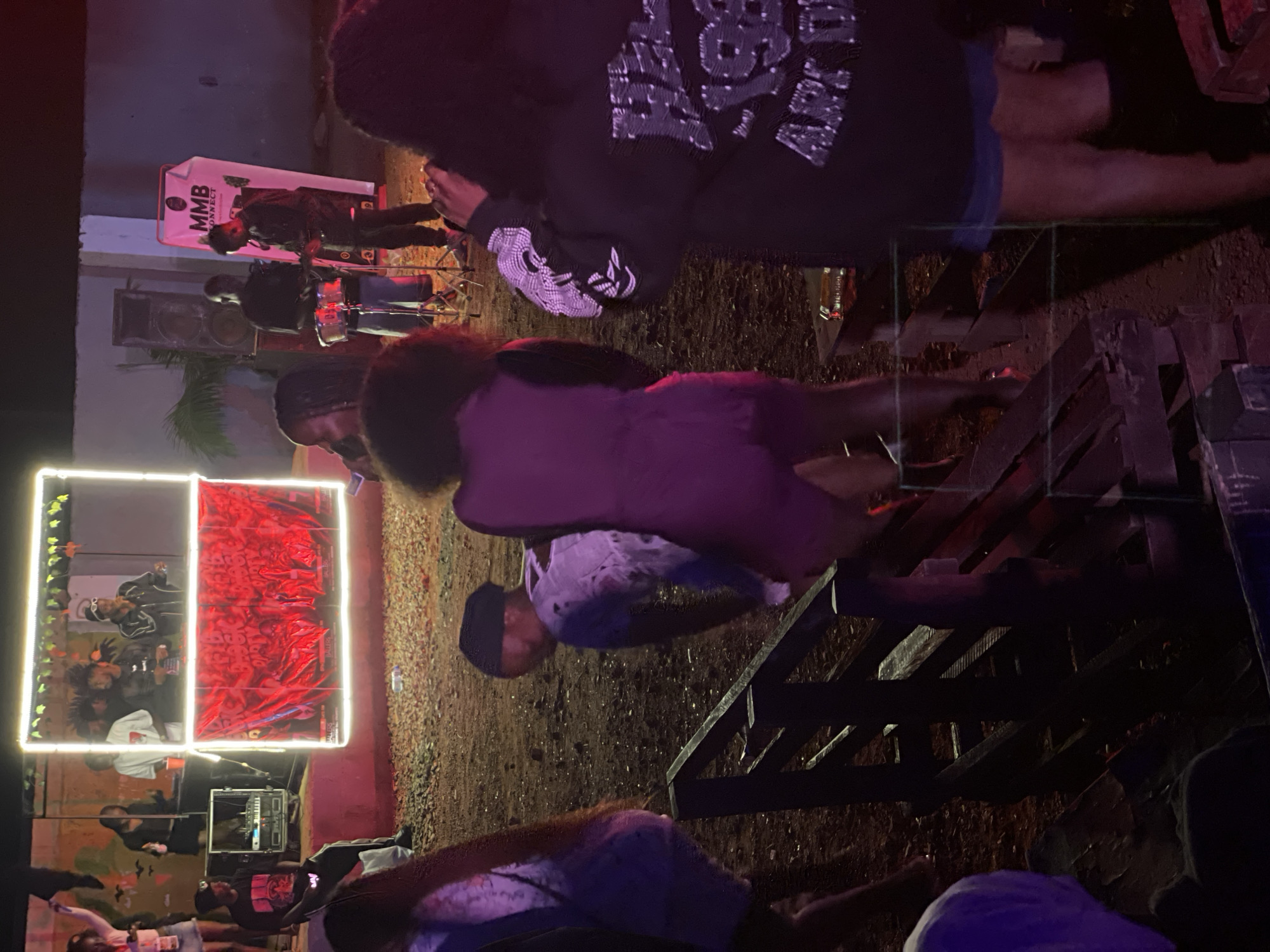 This image captures an outdoor gathering at night, likely in the city of Durumi, Nigeria. The scene is illuminated by neon lights surrounding a stage area, where several individuals are present. Two people are on stage, one appearing to be a performer speaking into a microphone, and another man standing beside them. Behind them, a large banner with red graphics and the text "BALLERS BLOCK PARTY" is displayed. To the left of the stage, a person is seated near DJ equipment. In the foreground and mid-ground, several other individuals are visible, some standing and others walking. The ground appears to be dirt or gravel, and there are wooden structures, possibly benches or seating areas. The overall atmosphere suggests a party or event.