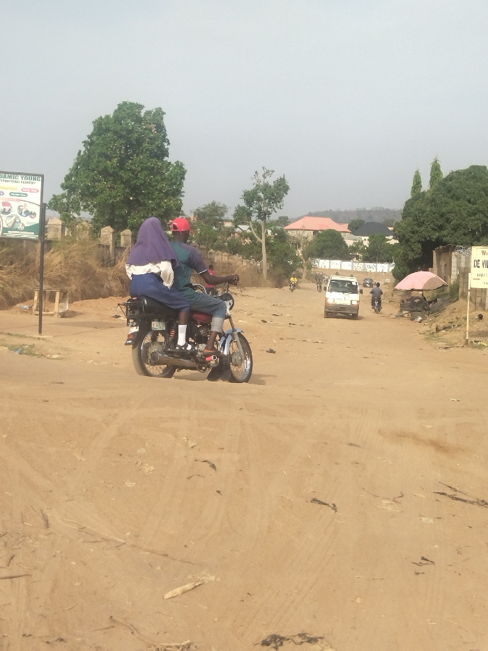 A man and a woman are riding a motorcycle on a dirt road in New Karu, Nigeria. The woman is wearing a blue hijab and the man is wearing a red hat. There is a white van in the distance and a few other vehicles are parked along the road. There are trees and bushes on both sides of the road. The ground is dusty and there are tire tracks visible. The sky is blue and there are some clouds in the distance. The road is empty except for the motorcycle. The scene is typical of a rural area in Nigeria. The image was taken in the afternoon, with the sun shining from above. The mood of the image is relaxed and peaceful.