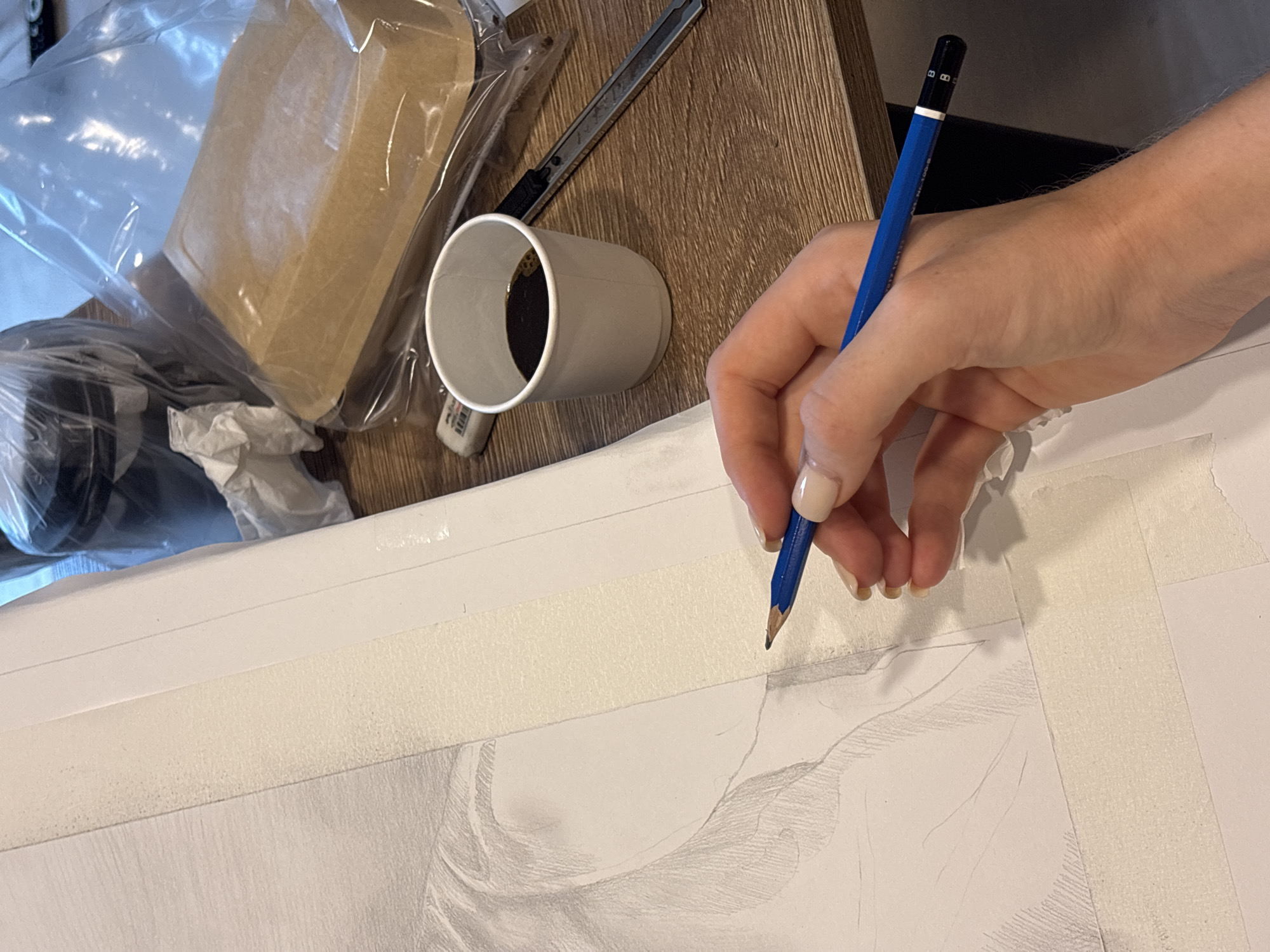 The image presents an overhead view of a person's right hand drawing with a pencil on white paper, set on a wooden surface. The scene depicts an indoor creative workspace, likely in Choeng Thale, Thailand.
The focal point is a fair-skinned hand, adorned with light-colored fingernail polish, holding a blue pencil with a black top. The pencil, which has "HB", "No. 1500", and "KOH-I-NOOR" faintly printed on its barrel and "HB" repeated on its black end cap, is sharpened to a fine point and actively engaged in sketching. The drawing, partially visible on the white paper, appears to be a graphite sketch, possibly a portrait or figure, characterized by soft outlines and initial shading. The paper is secured to the surface with light-colored masking tape, suggesting a stable setup for detailed work.
To the left of the drawing area, a white paper cup, containing a dark liquid presumed to be coffee or tea, rests on the wooden surface. Beside it, a small white eraser, faintly branded "KOH-I-NOOR", is visible. Further back on the wooden surface, which features a prominent grain, a silver utility knife or cutter with a black handle is placed. A brown paper bag, likely containing food, is enclosed within a clear plastic bag, with other clear plastic materials also scattered nearby, possibly packaging or waste.
The activity clearly shows the process of artistic creation. The indoor setting provides no specific time of day or weather cues, and no distinct emotions are discernible, but the arrangement of tools and materials suggests a focused and dedicated creative environment.