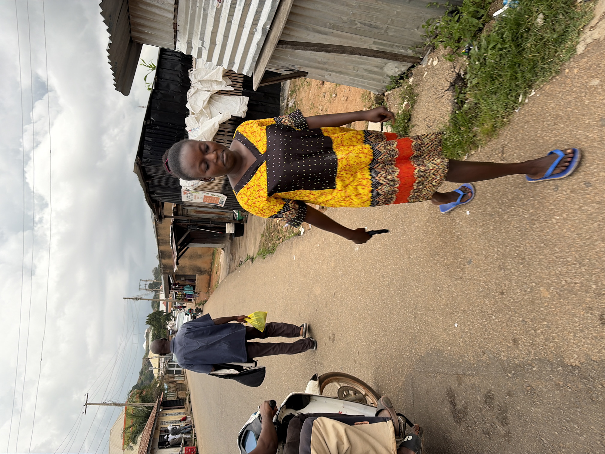 A woman in a yellow and black dress walks along a paved road in a street scene in Jos, Nigeria. She is wearing blue flip-flops and holding a black object in her right hand. To her left, a man in a blue shirt and brown pants walks away from the camera, carrying a black bag over his left shoulder and a yellow plastic bag in his right hand. A white motorcycle with a rider is parked on the left side of the road. Buildings with corrugated metal roofs line the right side of the road, with some showing exposed construction materials. A cloudy sky looms overhead, and the general environment appears to be an urban or semi-urban setting with a mix of residential and commercial structures.
