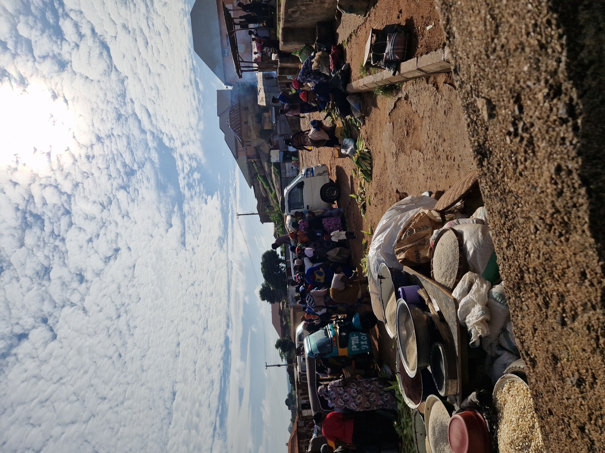 This image depicts a bustling outdoor market scene in New Stadium, Nigeria. The foreground is dominated by piles of dry soil and gravel, with several large bowls and bags of grains and other produce visible. In the mid-ground, a crowd of people, predominantly women dressed in vibrant clothing, are gathered around a turquoise motorized tricycle. Some are interacting with each other, while others appear to be engaged in selling or buying goods. Several vehicles, including a white van, are parked nearby. The background reveals a collection of buildings, some appearing to be homes with corrugated iron roofs, and others more structured with darker roofs. The sky is filled with dramatic, white, fluffy clouds, with the sun shining through, creating a bright and somewhat hazy atmosphere. The overall impression is one of lively commerce and daily life in a Nigerian urban setting.