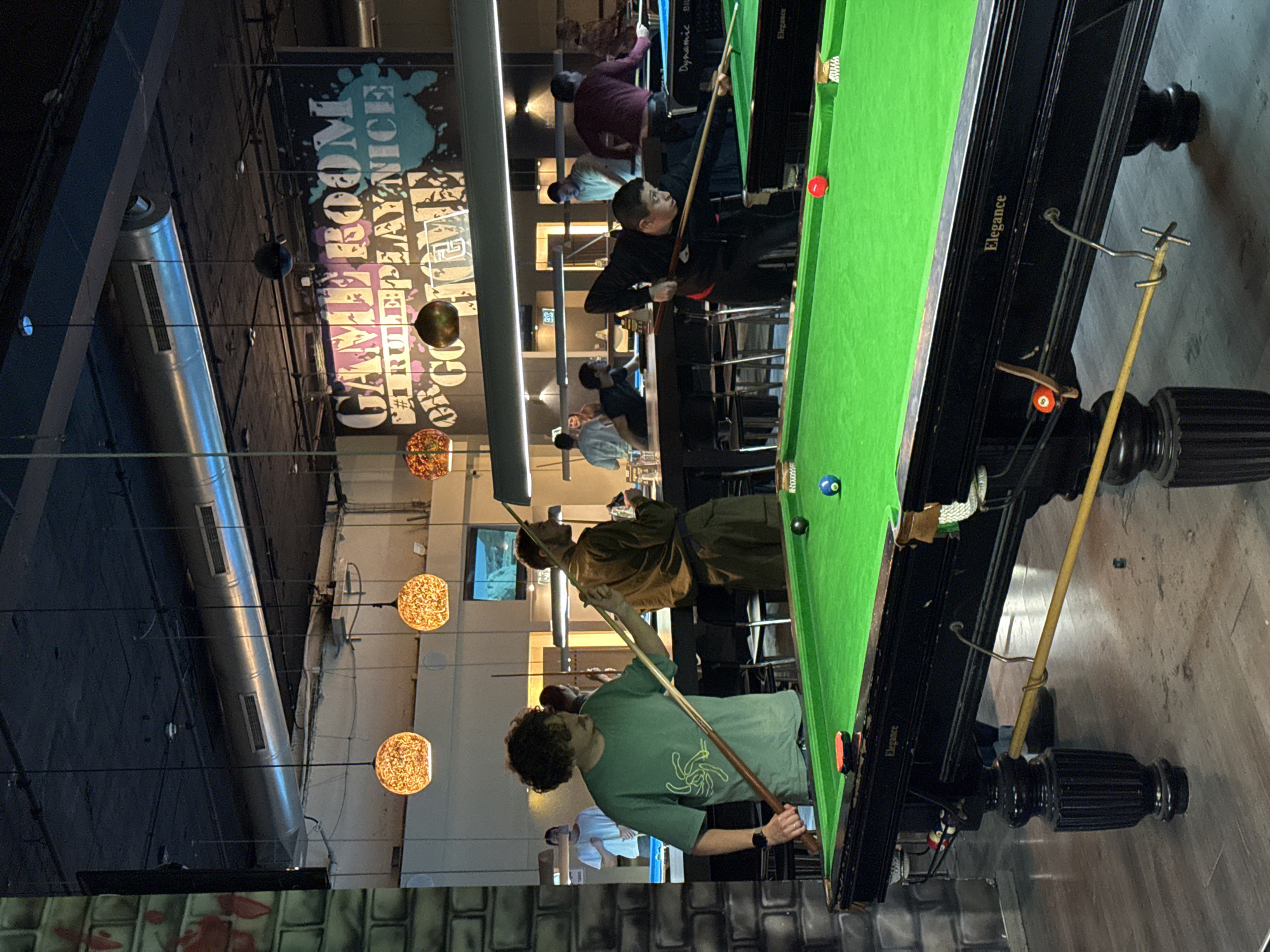 A brightly lit interior shows a game room scene. The focus is on several people playing pool at a table. A man wearing a green t-shirt is in the foreground, actively playing pool. Two men are standing by the table, observing the game. Further back, more people are playing pool at additional tables. Above the players, decorative, spherical light fixtures are suspended from the ceiling. Additionally, the wall displays the words "GAME ROOM #1 RULE PEOPLE DR GOODMAN" in a large, stylized font. The word “Elegance” is on the pool table. The location is likely in Tel-Aviv, Israel.