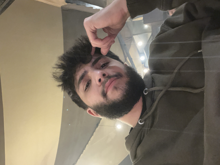 A bearded man with brown hair, wearing a green hooded sweatshirt, poses for the camera. He's holding his hand up, near his forehead, seemingly resting or scratching. The lighting appears artificial, with visible light fixtures in the background. The camera angle is slightly tilted, giving a casual, upward-looking perspective. The man's expression is calm, with a slight smirk.