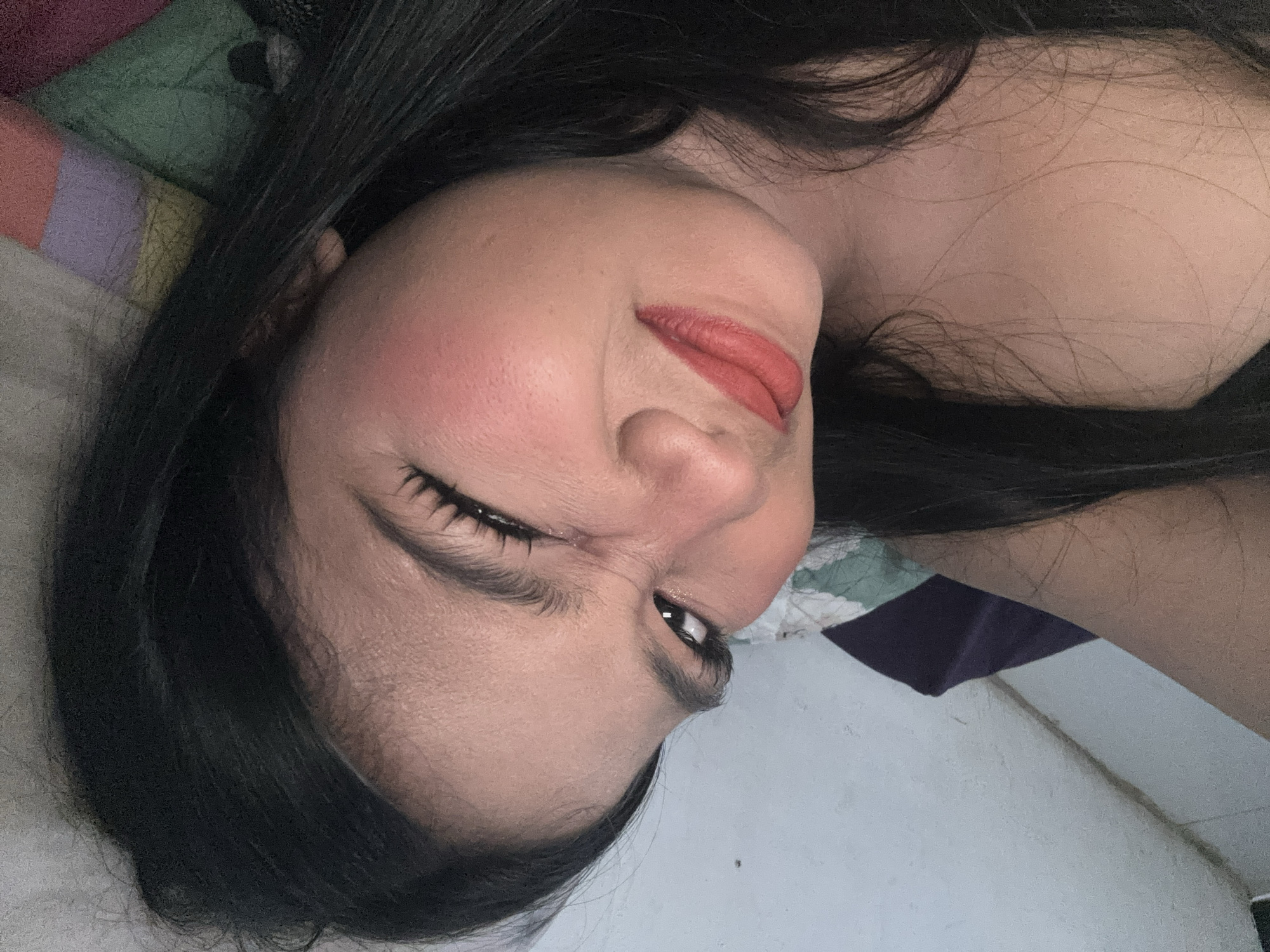 In this close-up, low-angle, portrait-oriented selfie, a young woman with long, dark hair is lying down. She is wearing makeup, including reddish-orange lipstick and blush. Her right eye is closed in a wink, while her left eye is open and looking towards the camera. Her facial expression appears playful. The background consists of a wall and bedding with a colorful striped pattern. Based on the lighting and the style of the bedding, it is plausible this image was taken indoors in a residential setting in North Jakarta, Indonesia.
