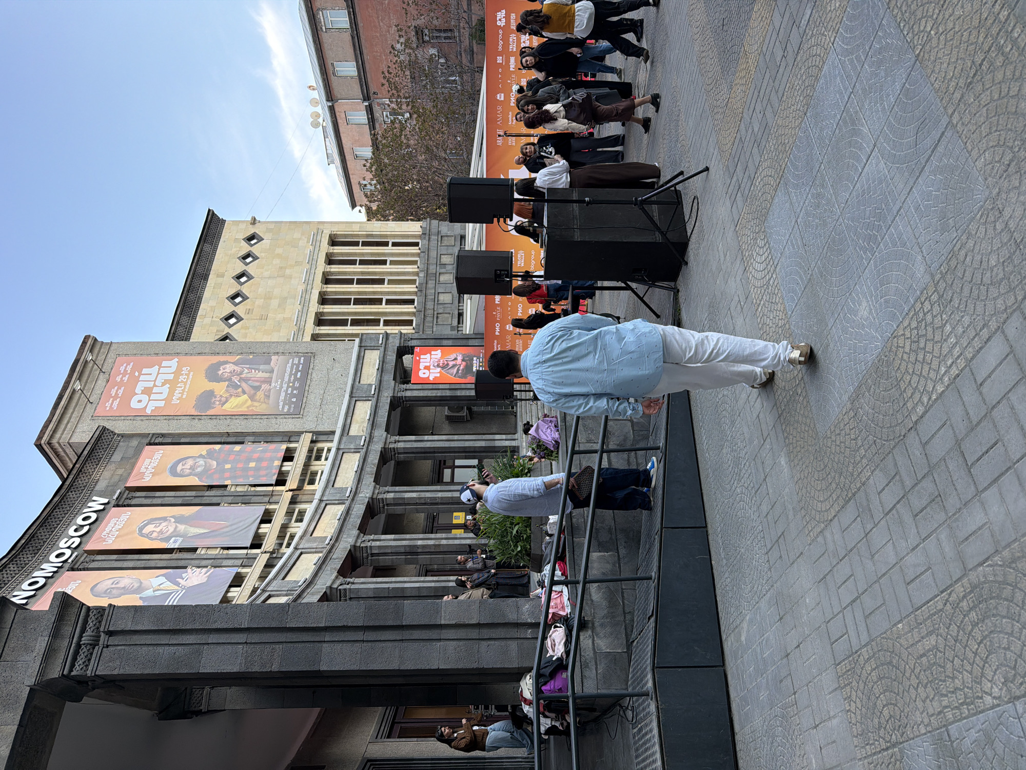 An outdoor urban scene in Yerevan, Armenia, depicts a lively public event on a clear day. The foreground features a tiled pavement with intricate grey and light yellow patterns. In the center, a man in a light blue shirt and white pants stands with his back to the camera, facing a temporary stage setup.
The stage area, positioned in the mid-ground, includes two large black speakers on stands. Behind them, a promotional backdrop displays various sponsor logos and text, including "TELCELL WALLET," "bbgroup," "AMAR," "PRIO," "PAVLE," "PRIME," and "Jellyfish." A group of people is gathered around the stage, some looking towards the unseen performers, others appearing to be involved with the event organization.
On the left, a large multi-story building with ornate architecture serves as a prominent backdrop. Its facade is adorned with several large banners. One banner partially visible on the far left reads "NOMOSCOW." Other banners feature portraits of individuals and Armenian text, including "ԱՊՐԻԼԻ 29-ԻՑ" (April 29th) and names like "ՆԱՀԱՊԵՏՅԱՆ," "ԿԱՐԱՊԵՏՅԱՆ," and "TILO," suggesting a cultural event, possibly a film festival or concert. People are also visible on a balcony or elevated walkway of this building, observing the activities below. In the far background, another building with windows and satellite dishes is visible against a bright blue sky. The overall atmosphere suggests a public performance or gathering.