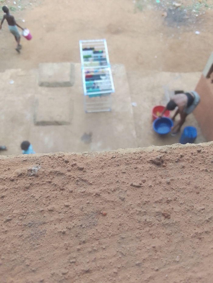 The scene depicts an outdoor ground area in Ukwu-Abwa, Nigeria, viewed from an elevated position, with a textured, granular orange-brown surface occupying the foreground. The ground is primarily exposed light brown dirt.
Three individuals are present. In the upper left, a shirtless individual in light-colored shorts walks rightward, carrying a pink plastic bucket in their left hand. To the right, another individual, wearing a dark top and light bottom, is bent over, facing left, interacting with three plastic basins on the ground: a red one, a blue one, and a lighter blue/white one. Further to the lower left, a third, heavily blurred individual wearing a blue top is partially visible.
Centrally located is a white metal clothes drying rack, a multi-tiered structure, with various multicolored garments hanging on its rungs. Two rectangular concrete slabs are embedded in the dirt ground to the left of the drying rack. A section of an orange-brown building wall is visible on the far right. The background shows more dirt ground with minimal, indistinct vegetation or debris.