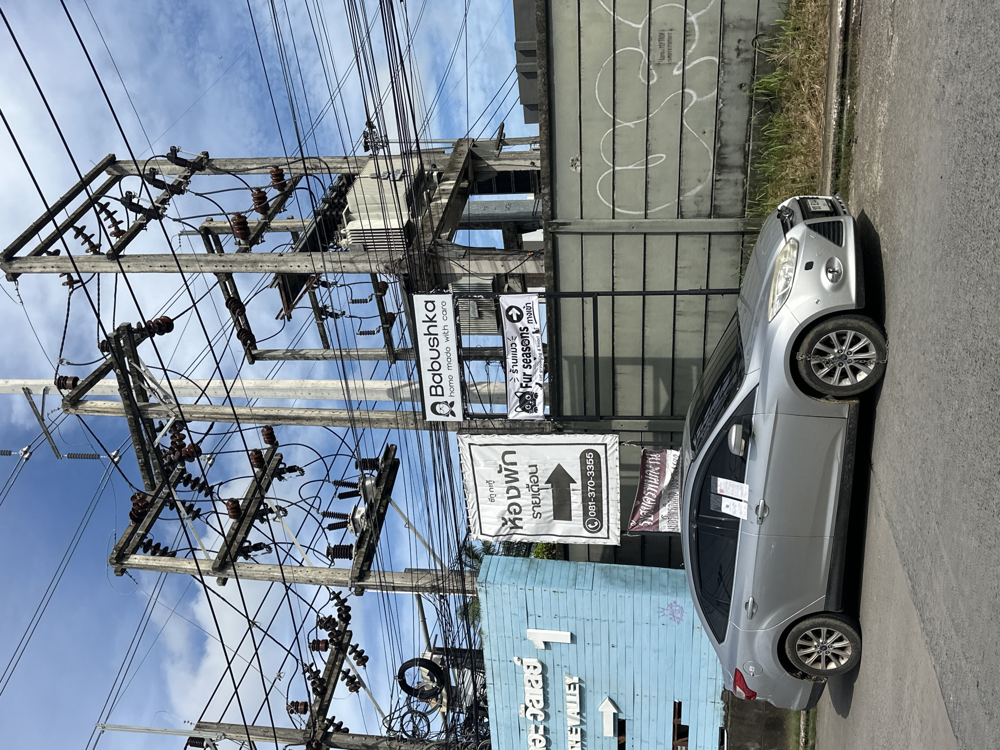 In Kathu, Thailand, a silver Ford Focus sedan is parked on a paved road next to a light blue building with the words "THE VALLEY" and the number "1" visible. A large utility pole with numerous electrical wires and transformers dominates the background. Several signs are attached to the utility pole. One sign, in black and white, reads "ห้องพัก รายเดือน" (rental rooms monthly) with an arrow pointing to the right and a phone number "081-370-3355". Another sign, with a cat graphic, says "Fur seasons" and below it "cat grooming & hotel". A third sign, partially obscured, displays the text "Babushka" and "home made with care". A concrete wall with graffiti runs behind the car and the utility pole. The sky above is partly cloudy. Two white notices are affixed to the driver's side windows of the car.
