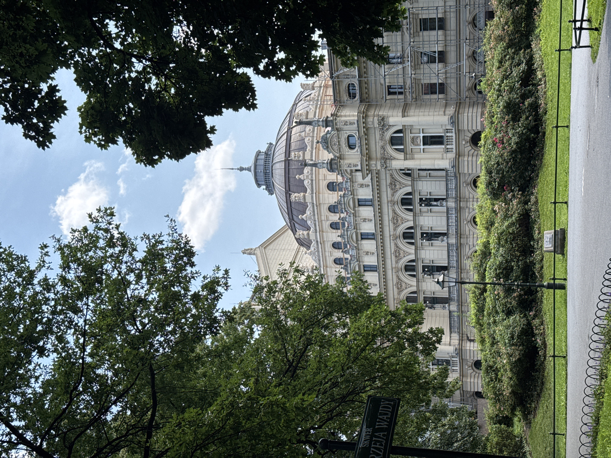 The image shows a partial view of the Juliusz Słowacki Theatre in Krakow, Poland, on a partially cloudy day. The theatre is a grand building with a domed roof, ornate facade, and classical architectural elements. It is surrounded by lush green trees and manicured bushes. In the foreground, a street sign with Polish text and a black metal fence can be seen. The scene suggests a calm, pleasant day in a park or public square, with the theatre as the prominent focal point.