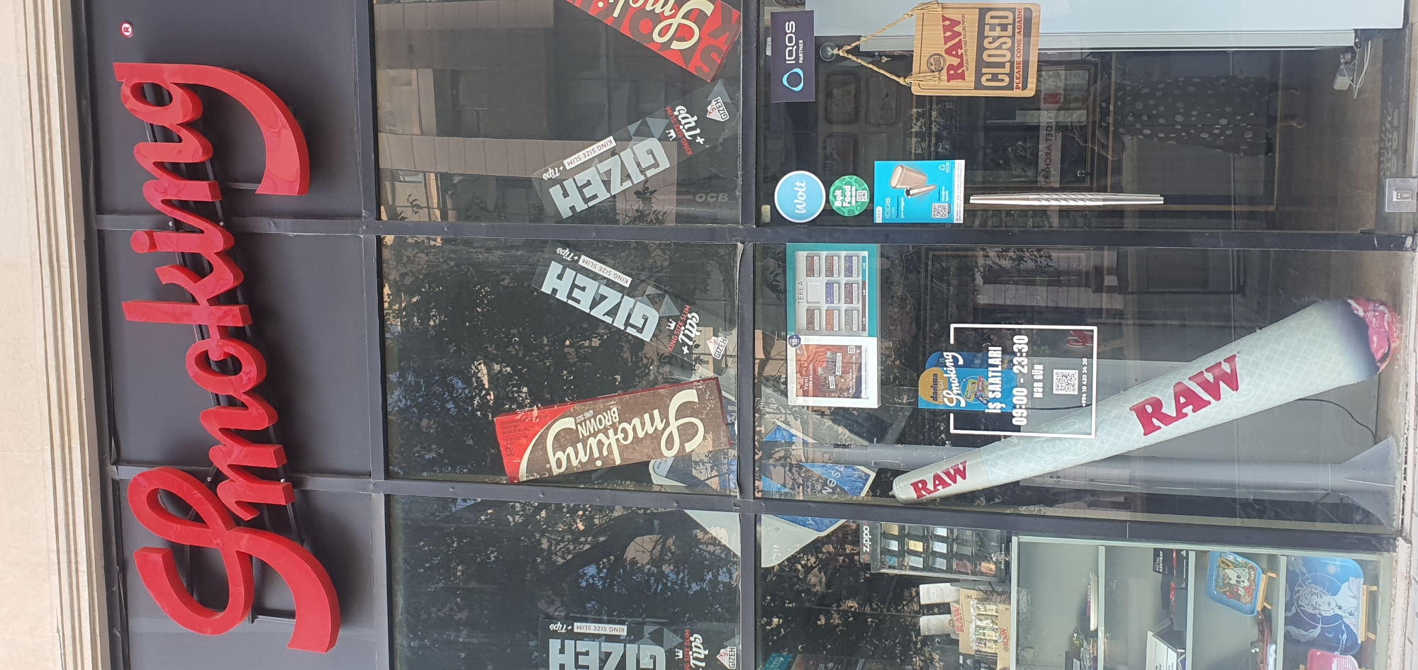 The image shows the storefront of a business named "Smoking" in Baku, Azerbaijan. The store has a large red script sign on a dark facade above a glass-paned storefront. The glass displays various products and advertisements, including branded rolling papers, lighters, and what appears to be a large, inflatable cigarette. A sign indicating "IS SAATLARI" (Working Hours) with hours from 09:00 to 23:30 is visible on the right-hand side of the window. Another sign at the entrance reads "CLOSED PLEASE COME AGAIN!". The reflection in the glass shows buildings and trees, suggesting an urban environment. A woman in a polka dot dress can be seen reflected in the glass, appearing to be taking a photograph.