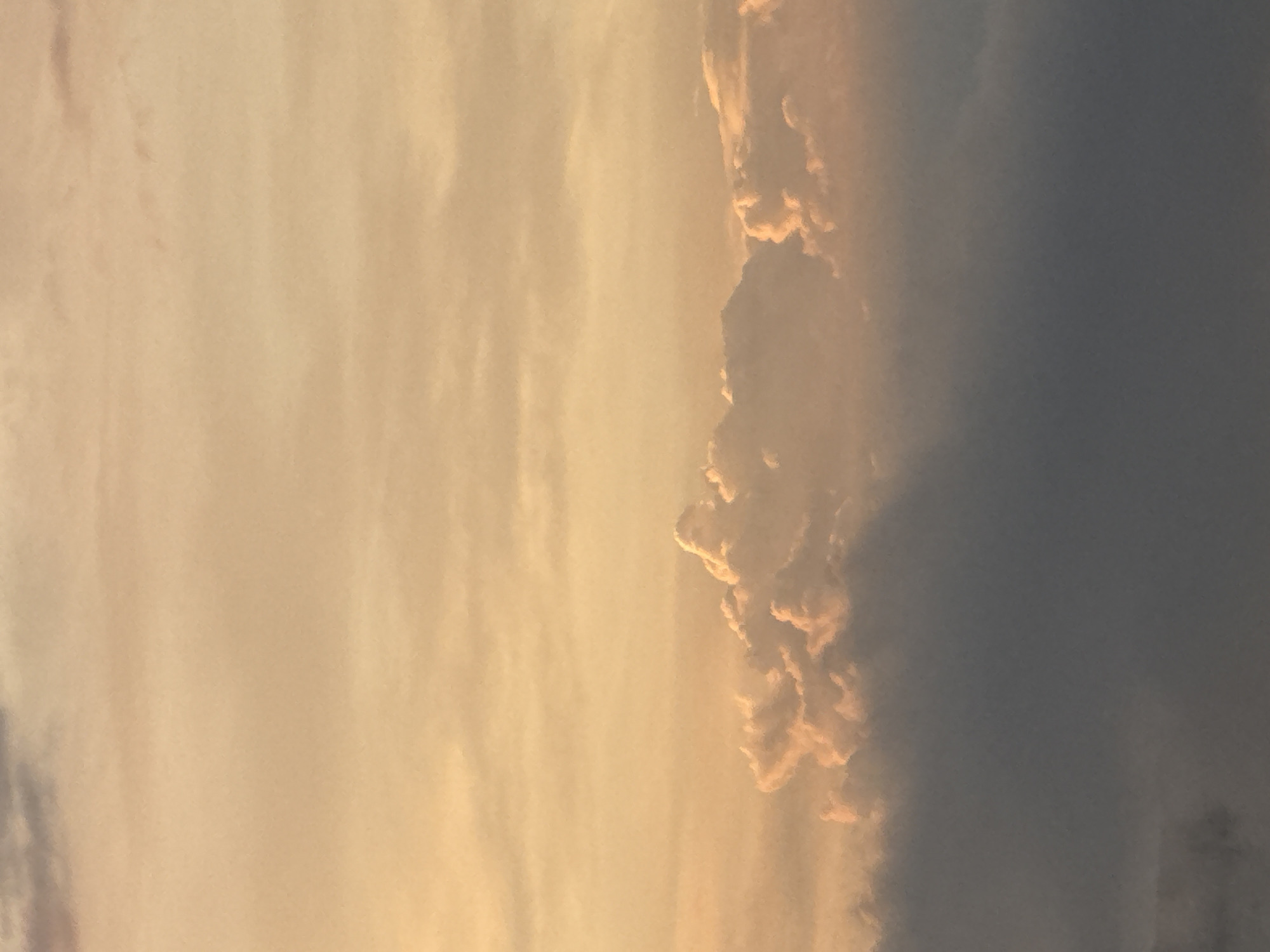 A vertical, full-frame, medium shot photograph captures a cloudy sky at sunset, illuminated by warm, golden light. The sky is a gradient of soft yellow and orange, with diffused light suggesting the sun is low on the horizon. Layered clouds are prominent; the lower layer is dark gray and dense, obscuring the view below, while the upper layer features lighter, more textured clouds tinged with pink and orange from the setting sun. The overall impression is one of atmospheric beauty and tranquility.