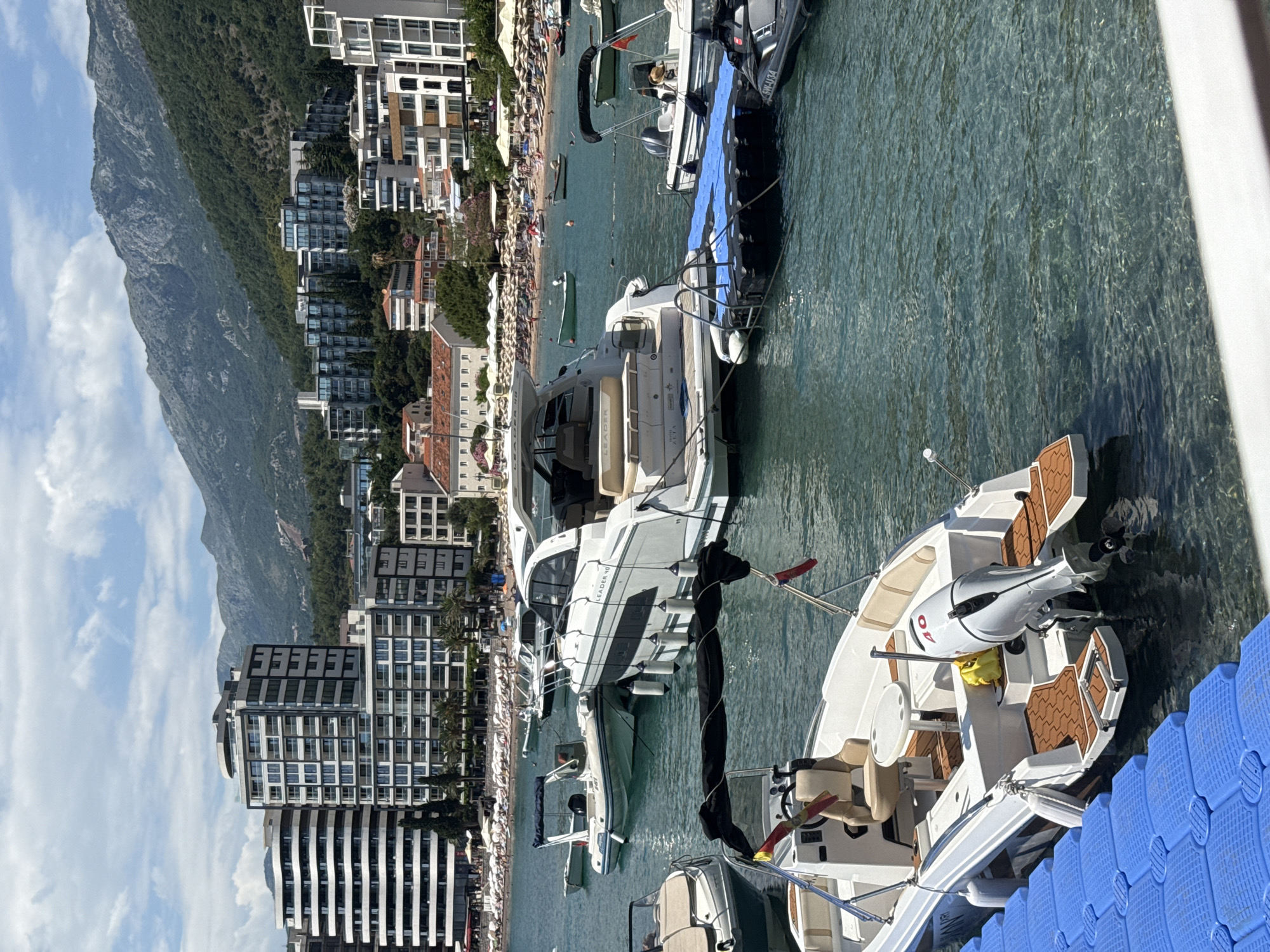 The image depicts a scenic view of a coastline in Rafailovići, Montenegro, characterized by a picturesque beach lined with numerous sun umbrellas and beach chairs, with a scattering of people enjoying the seaside. Behind the beach, modern, multi-story buildings and some older structures form a dense urban landscape that stretches along the coast. The buildings vary in architectural style, from sleek, contemporary designs with large windows to more traditional facades with red-tiled roofs.
In the foreground and middle ground, several boats are moored in clear, blue-green water. These include a large white motor yacht with the text "LEADER 40" visible on its hull, and other smaller vessels, some of which appear to be speedboats or tenders. A dark blue floating dock is visible to the right of the frame, connected to the boats. The water is calm and transparent, allowing a view of the seabed in some areas.
Towering over the coastal town and the water is a large, lush green mountain. The mountain's slopes are densely covered with trees, and its rocky terrain is visible in places. The sky is partly cloudy with scattered white clouds against a bright blue backdrop, suggesting pleasant weather, likely during the daytime, possibly mid-morning or early afternoon. The overall impression is one of a vibrant tourist destination, combining natural beauty with urban development and recreational activities on the water.