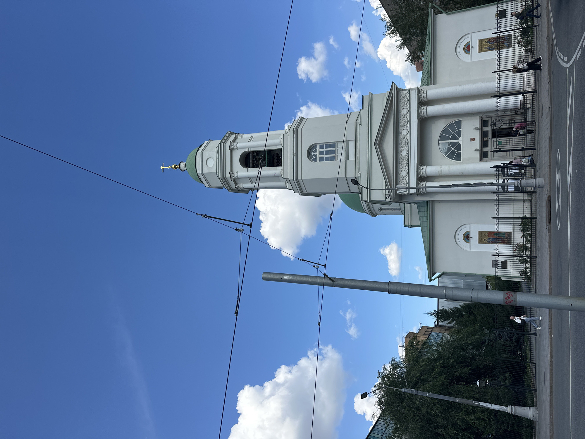 The image shows a view of the Church of the Assumption of the Virgin Mary at Pokrovka in Moscow, Russia. The white church features a prominent bell tower with a green dome and a golden cross. The building is adorned with classical columns and arched windows. In front of the church, a black metal fence encloses a small garden. Several people are visible walking near the entrance of the church. The sky is blue with scattered white clouds, and utility wires are strung across the frame. A street and sidewalk are in the foreground, with a lamppost and trees to the left.