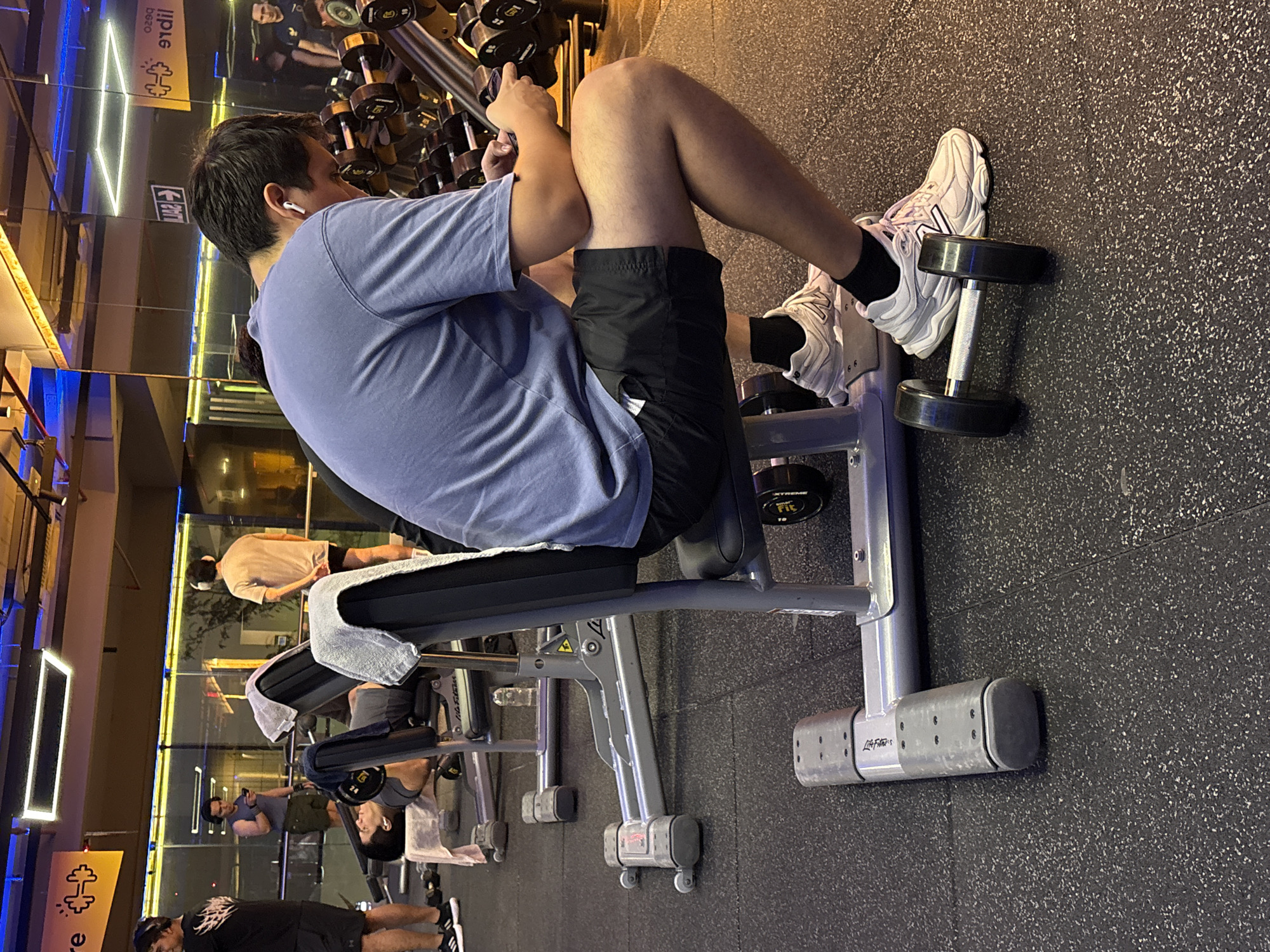 In a gym in San Isidro, Peru, a man wearing a blue t-shirt and black shorts sits on a weight bench. He is wearing white wireless earbuds and holding a mobile phone in his right hand. A dumbbell rests on the floor in front of him, and other dumbbells are visible on a rack in the background. The gym floor is black rubberized material. The scene captures a moment of rest or engagement with a device amidst a workout setting.
