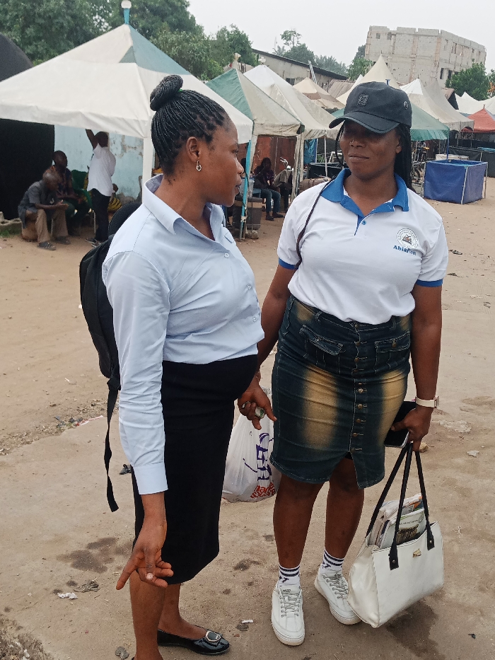 Two adult females are standing and conversing outdoors in Ura Nta Umuarandu, Nigeria. The ground is an uneven, light brown dirt surface.
The female on the left wears a light blue long-sleeved collared shirt, a black pencil skirt, and black flat shoes. Her hair is braided and styled into a bun. She wears a small hoop earring in her visible ear and carries a black backpack on her left shoulder. She holds a white plastic bag in her right hand and points her left hand downwards.
The female on the right wears a black baseball cap, a white polo shirt with blue trim and an "AbiaFm" logo on the left chest, a faded blue and yellow-brown denim midi skirt, white athletic sneakers, and white socks with blue stripes. She wears a light-colored wrist watch on her left wrist. She holds a white tote bag containing papers in her left hand and a dark-colored smartphone in her right hand.
Both women are facing each other, engaged in interaction.
In the background, numerous pointed tents, primarily white with blue or green stripes, are erected, suggesting an outdoor market or event. Under a tent to the far left, several men are seated. To the right, a multi-story concrete building with multiple windows is visible. A blue rectangular container is also present in the mid-ground. The sky is overcast.