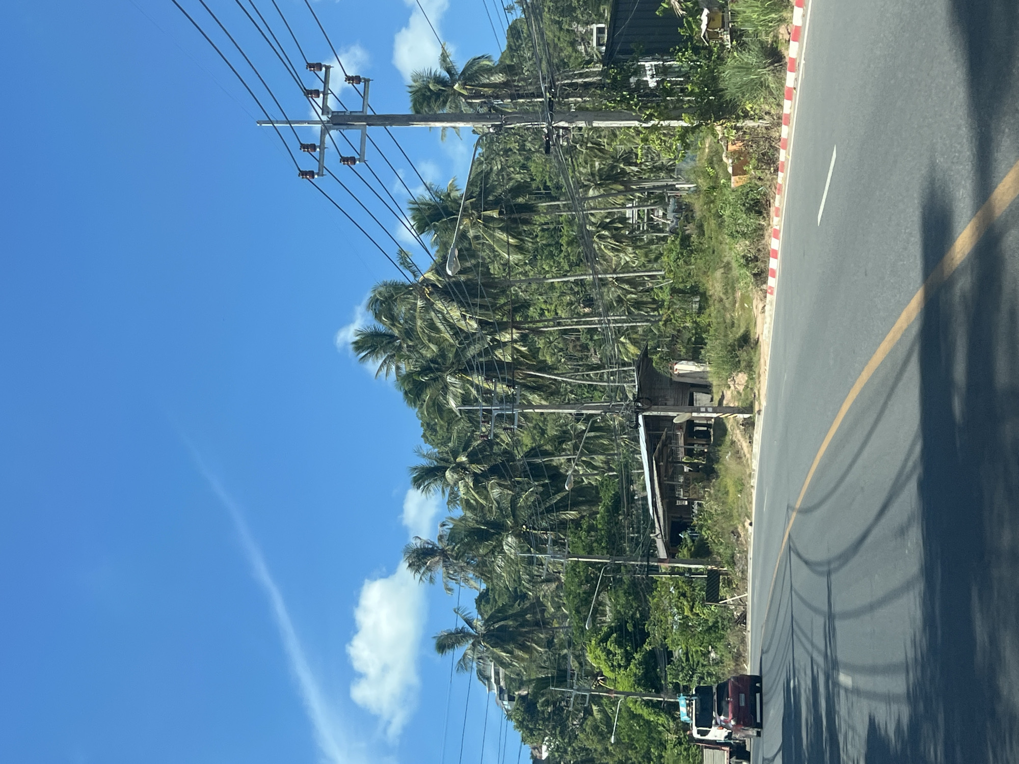 The image shows a road in Baan Chaweng Noi, Thailand, with a lush, tree-covered hillside in the background. The sky is a vibrant blue with a few wispy clouds. Coconut palm trees dominate the vegetation on the hillside, with some buildings and power lines visible among them.
In the foreground, the asphalt road has yellow and white lane markings. Tire tracks are visible on the road surface. A red car is driving on the road, followed by a white truck. The roadside is lined with red and white painted curbs and dense green vegetation. A small, rustic wooden structure with a corrugated roof is situated near the roadside. The overall impression is a tropical, somewhat undeveloped area, likely a coastal region given the typical scenery associated with Thailand.