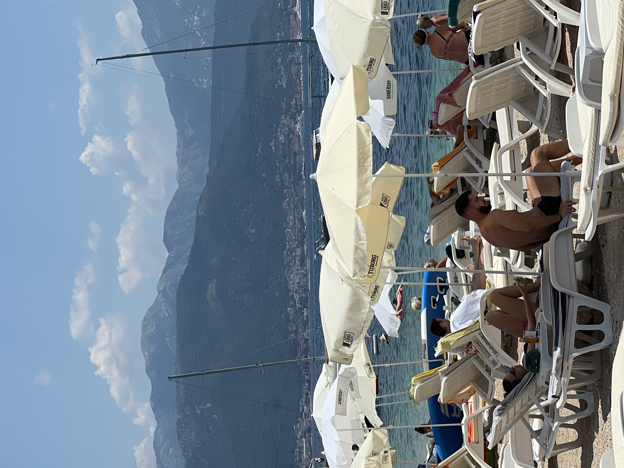 A busy beach scene unfolds in Baošići, Montenegro, under a clear blue sky dotted with fluffy white clouds. Numerous white and cream-colored beach umbrellas provide shade for lounge chairs occupied by people relaxing on the sand. The water is calm and appears to be a vibrant turquoise. In the background, sailboats with tall masts are moored, and further out, a town with buildings lines the coast. Beyond the town, a range of imposing, blue-toned mountains forms the horizon.
In the foreground, several individuals are visible. A woman in pink bikini bottoms and a black top sits on a lounge chair, holding a drink and a phone. Beside her, a man with dark hair and shorts is seated, looking away. Further back, another man reclines on a lounge chair. A woman in a bikini is seen in the water, floating on a pink inflatable. Other beachgoers are scattered across the scene, some lying down, others conversing. The overall impression is one of a lively and pleasant summer day at the beach.