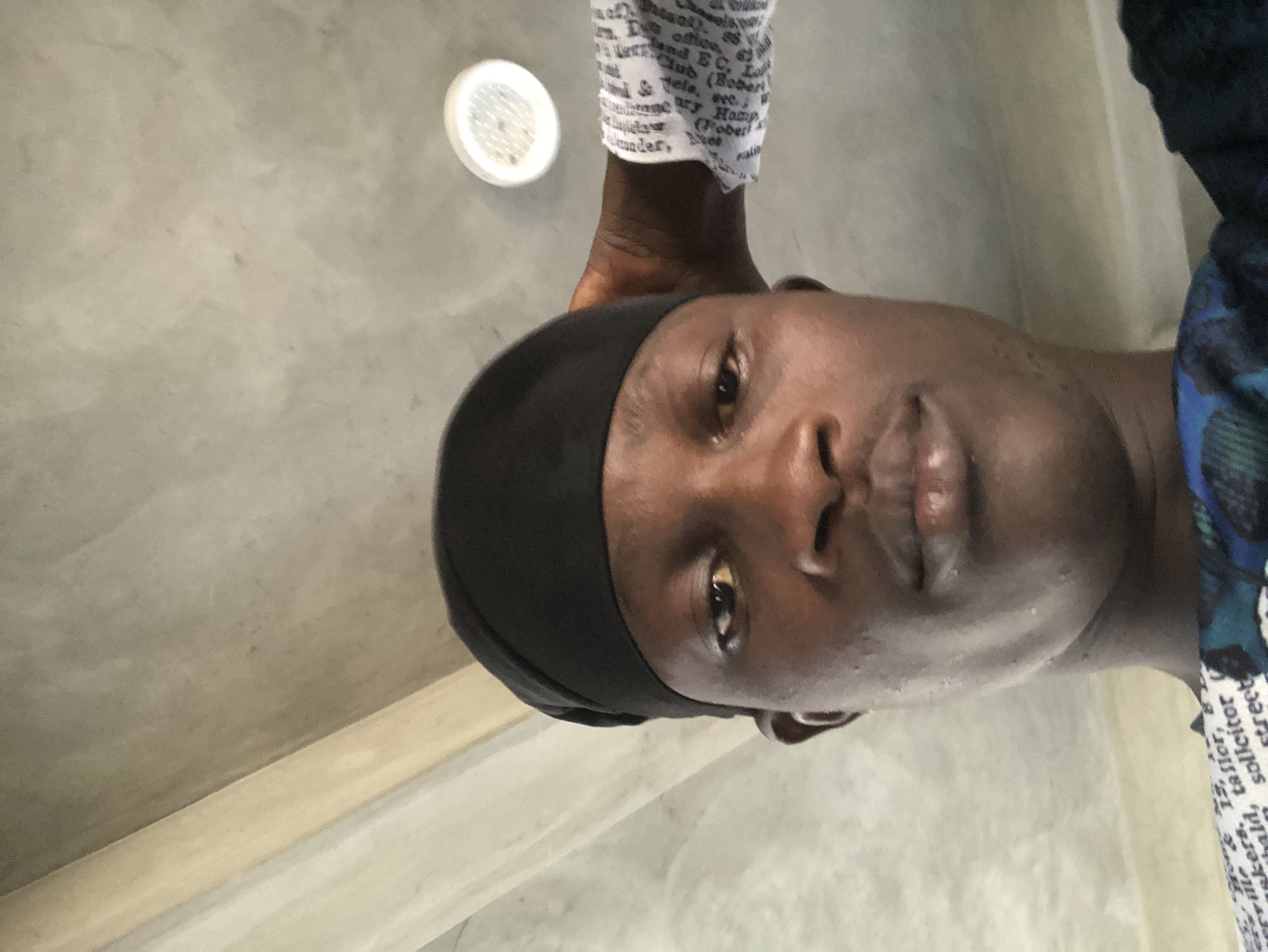 A close-up, overhead selfie shows a Black man wearing a black durag and a blue and green patterned garment. He is looking directly at the camera with a neutral expression. His right hand is visible, resting on the back of his head. The background consists of a textured ceiling with visible support beams and a light fixture. The image appears to have been taken indoors, possibly in a residential or commercial building. There is no discernible information in the image that would indicate the specific location within Ado, Nigeria.