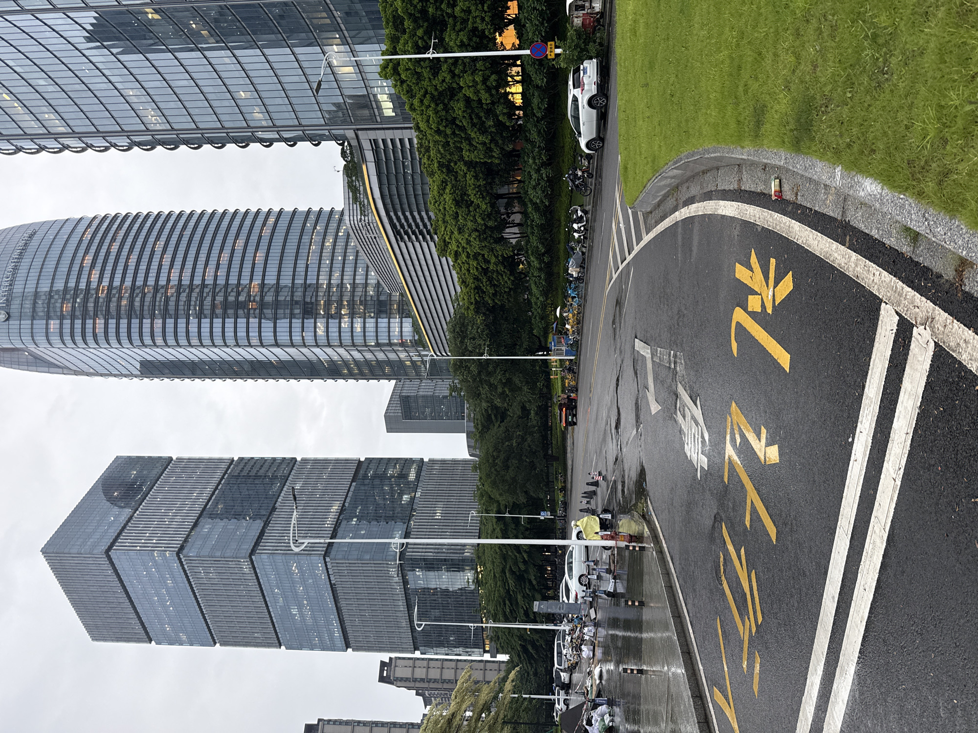 The image depicts an urban scene with modern high-rise buildings, a street, and a grassy area. The buildings are characterized by their glass facades and intricate architectural designs, with some reflecting the overcast sky. In the foreground, a wet asphalt road features yellow painted markings, including arrows and text, suggesting a traffic or directional cue. Several vehicles, including cars and numerous parked motorcycles and electric scooters, are visible along the roadside. A line of trees separates the road from the buildings. The presence of parked vehicles and the wet road surface indicate that it may have recently rained or is currently raining, suggesting a cloudy or damp weather condition. The scene appears to be a city environment, likely in a commercial or business district, given the presence of large office buildings and a variety of transportation options. The overall mood is neutral, presenting a typical urban landscape. The visible text in yellow on the road is likely in Chinese characters, possibly indicating a parking zone or traffic instruction. The location context specifies Haizhu District, China.