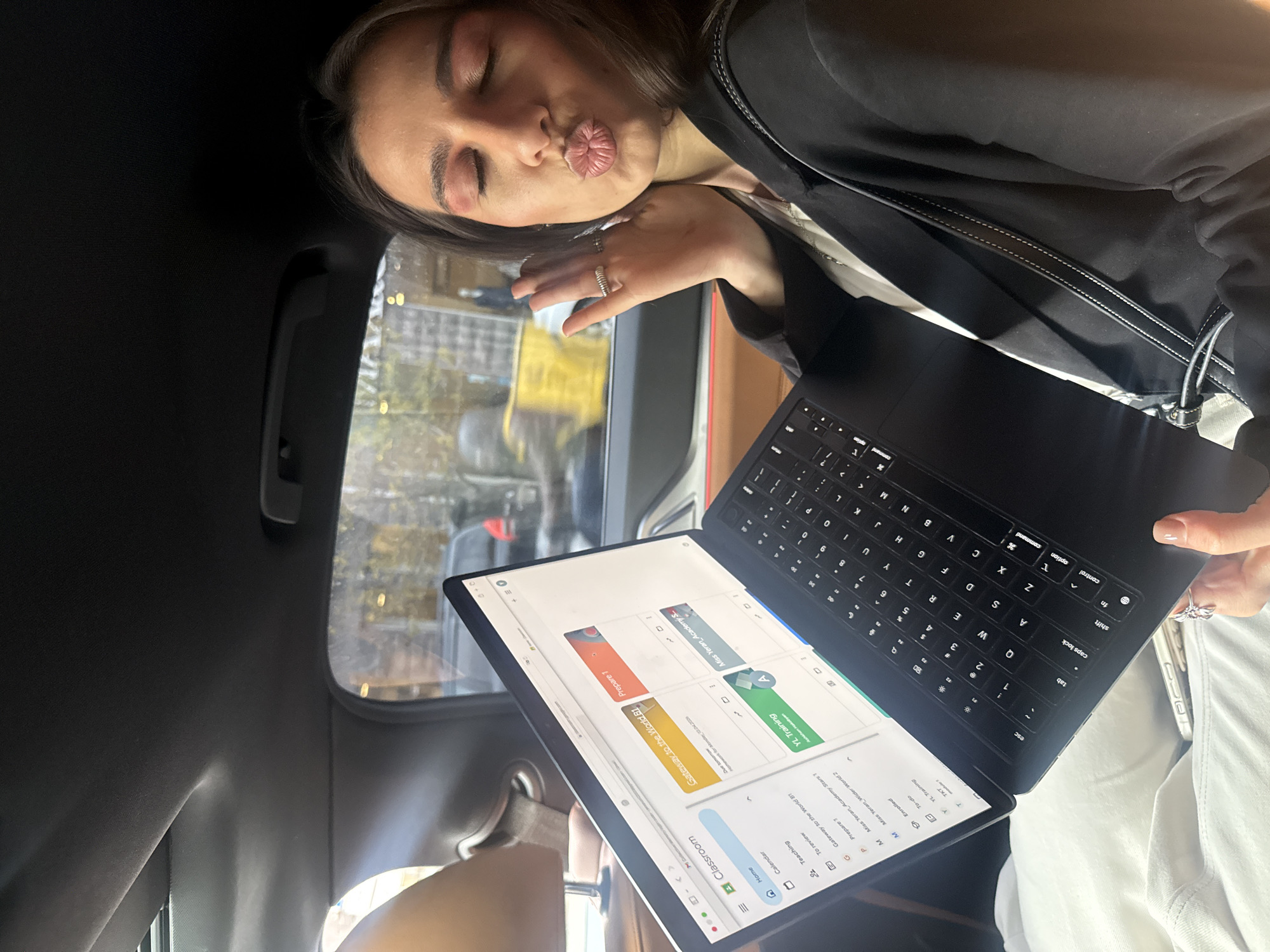 A woman is posing for a photo inside a car. She's holding a black laptop open to a screen displaying Google Classroom with the title "Gateway to the World B1" and the name "Miss Yeran Academy Stars." She's wearing a black jacket, and her lips are puckered in a kiss. Through the car window, blurry street scenes suggest an urban area. Based on the screen content, the location is likely in Yerevan, Armenia.