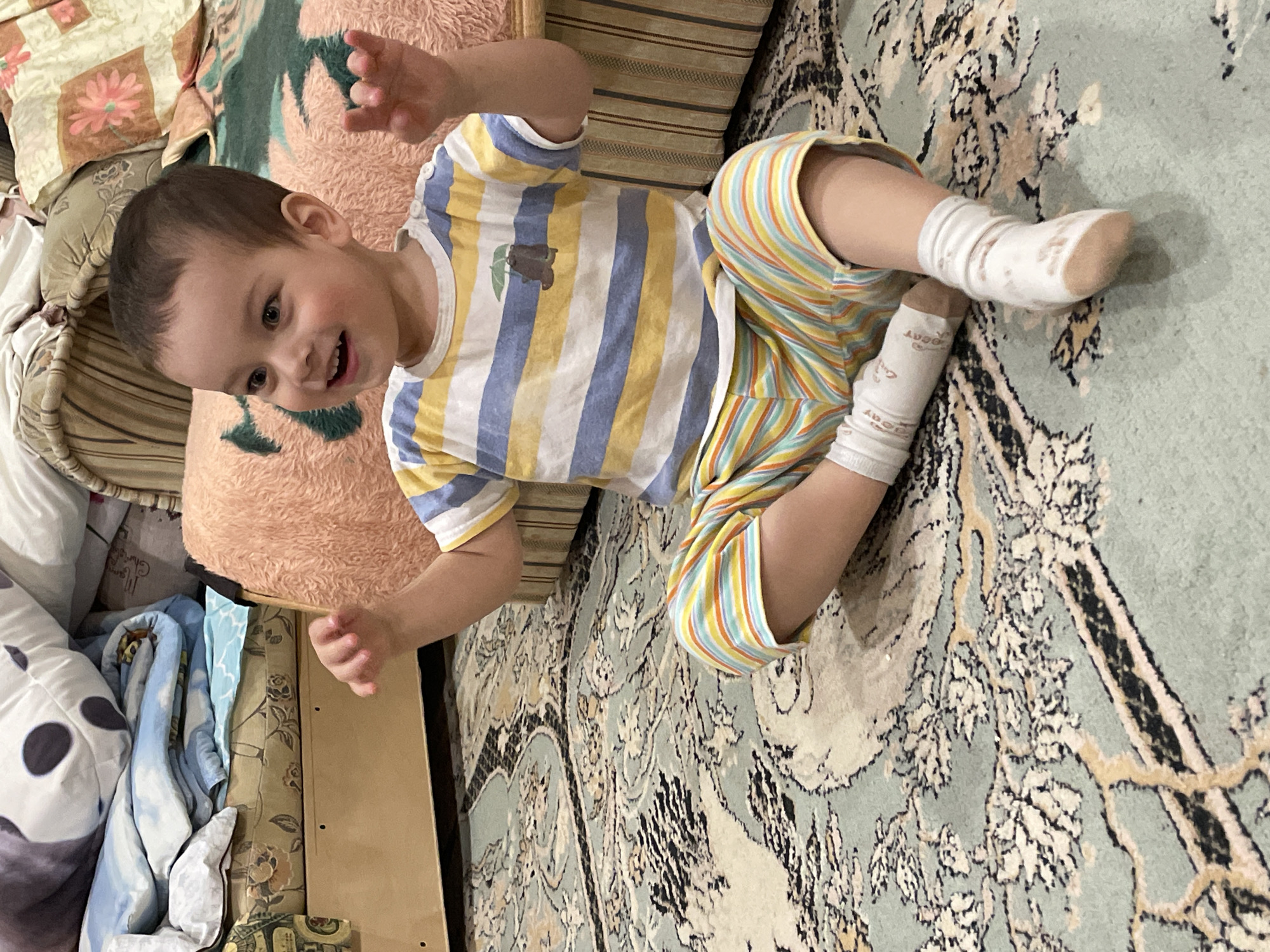 A young child lies on an ornate rug, smiling and holding their hands up in the air. The child wears a striped shirt and striped shorts with white socks. The rug is predominantly light blue and patterned with floral motifs in black and brown. Behind the child are various pillows and bedding, including a peach-colored, shaggy pillow. A wooden furniture piece is visible on the left side of the frame, near the bedding. The location is Zav'yalovo, Russia.