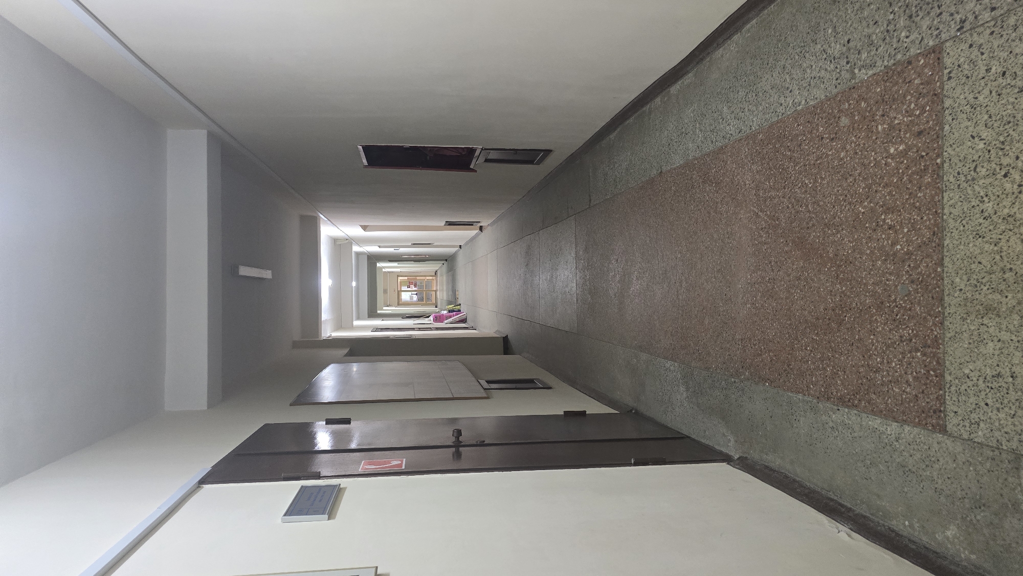 This media file shows a long, brightly lit corridor looking down from a high angle, giving a sense of extreme perspective. The corridor appears to be in a building with multiple floors, as suggested by the receding openings in the ceiling and the repeating pattern of doors and walls. The walls are a neutral color, and the floor is covered in a speckled terrazzo material, with a large rectangular inlay of reddish-brown terrazzo in the center.
The corridor is sparsely furnished. In the foreground, there are several dark brown doors, one of which has a red and white fire exit sign visible. There are also some flat, dark surfaces that could be tables or shelves. Further down the corridor, near the center, are a few items that appear to be stacked luggage or bags, in shades of pink and white. At the far end of the corridor, a set of glass doors leads to another section of the building, which is also illuminated.
The overall impression is one of institutional architecture, possibly a dormitory, apartment building, or hospital. The lighting is bright and even, suggesting it is daytime. There is no visible weather, as the scene is entirely indoors. The extreme perspective, with the corridor narrowing to a single point in the distance, creates a dramatic visual effect. The scene is devoid of people, contributing to a sense of stillness and perhaps slight emptiness.
Some text is visible: a red and white fire exit sign on a door in the foreground. Another small, rectangular sign with white text on a blue background is visible on a white surface in the foreground.