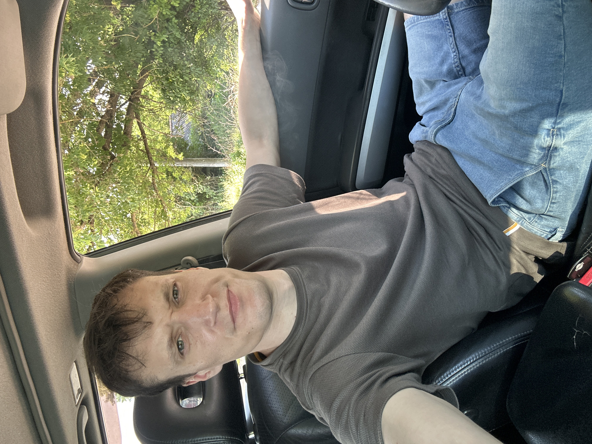 A man with short brown hair and green eyes is sitting in the driver's seat of a car, with his right arm resting on the open car door. He is wearing a dark grey t-shirt and blue jeans. The interior of the car is visible, including a black leather seat and the car's dashboard. Outside the car, green trees and foliage are visible, illuminated by sunlight. The scene appears to be in a rural or suburban area, as indicated by the presence of trees and a building in the background. The location of Makiv, Ukraine is noted.