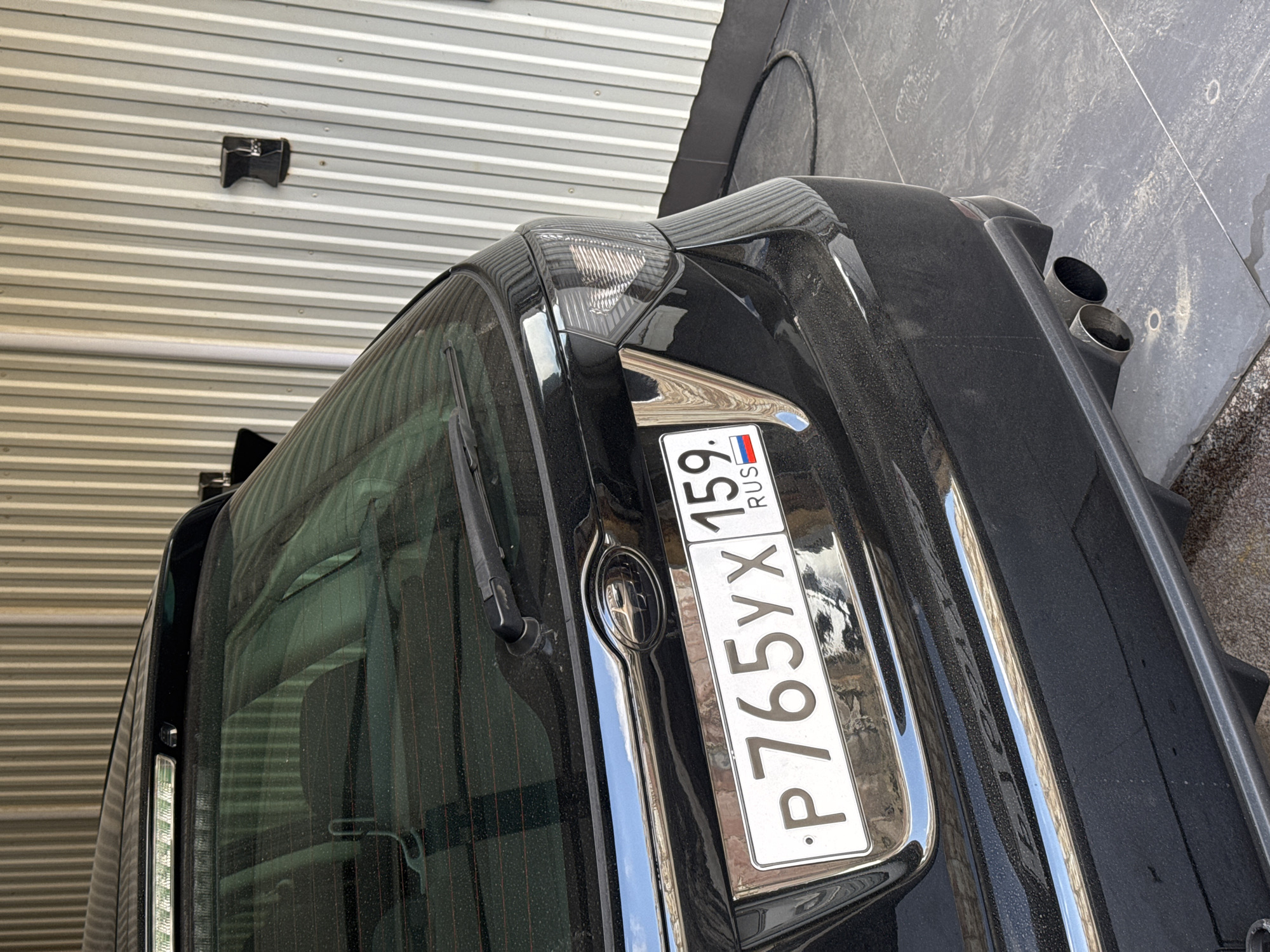 The image shows the rear of a black car with a Russian license plate. The license plate reads "P765YX 159 RUS". The car is parked next to a wall with horizontal metal panels and what appears to be a gray concrete or stone surface. The car has dual exhaust pipes visible on the right. The location is identified as Chaikovsky, Russia based on the license plate region code (159).