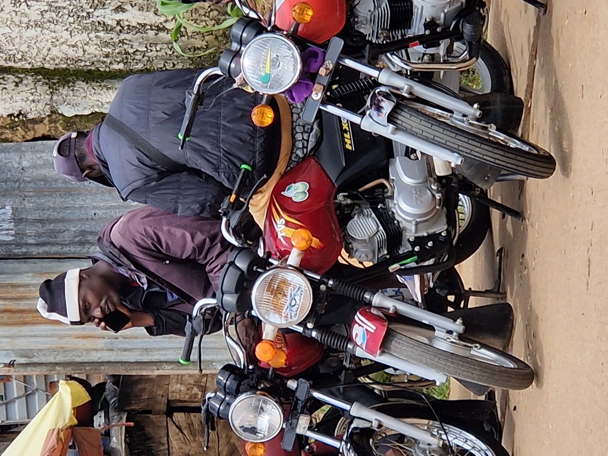 Two men are seated on motorcycles in Kabong, Nigeria. The man on the left, wearing a black and white striped beanie and a maroon jacket, is holding a black smartphone to his ear. The man on the right, wearing a dark cap and a black puffer vest over a maroon shirt, is facing away from the camera. Three motorcycles are visible, two in the foreground and one partially obscured behind them. The motorcycles are red and black. The background consists of a corrugated metal wall and some vegetation. The ground is dusty.