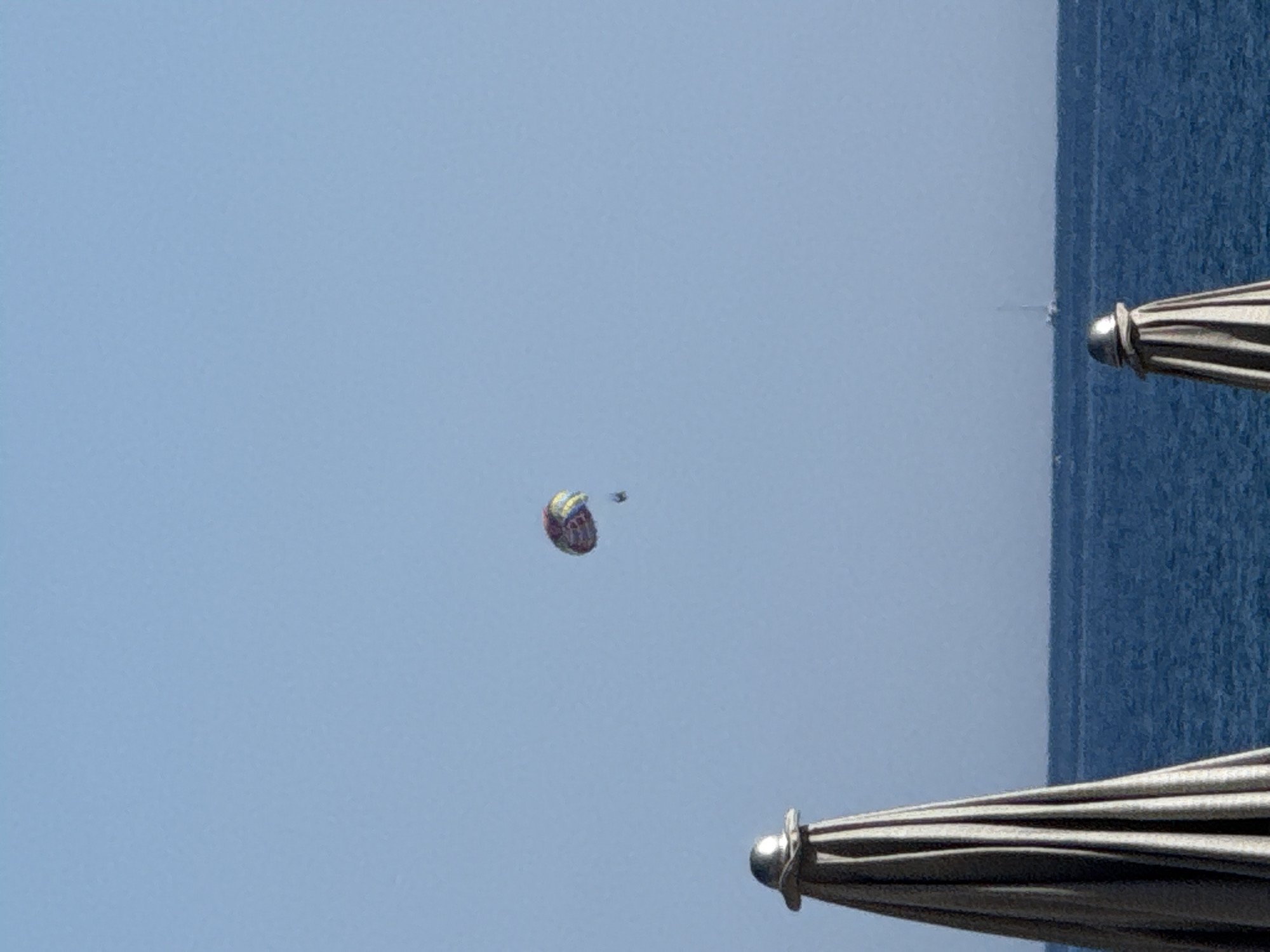 A parasailer is in the sky above the Adriatic Sea off the coast of Rafailovići, Montenegro. The parasailer is visible as a small, colorful canopy with two people beneath it, suspended in the blue sky. In the foreground, two beach umbrellas, folded and covered, are positioned to the left and right of the frame. The sea is a deep blue, and a small sailboat is visible on the horizon. The sky is clear and bright blue.