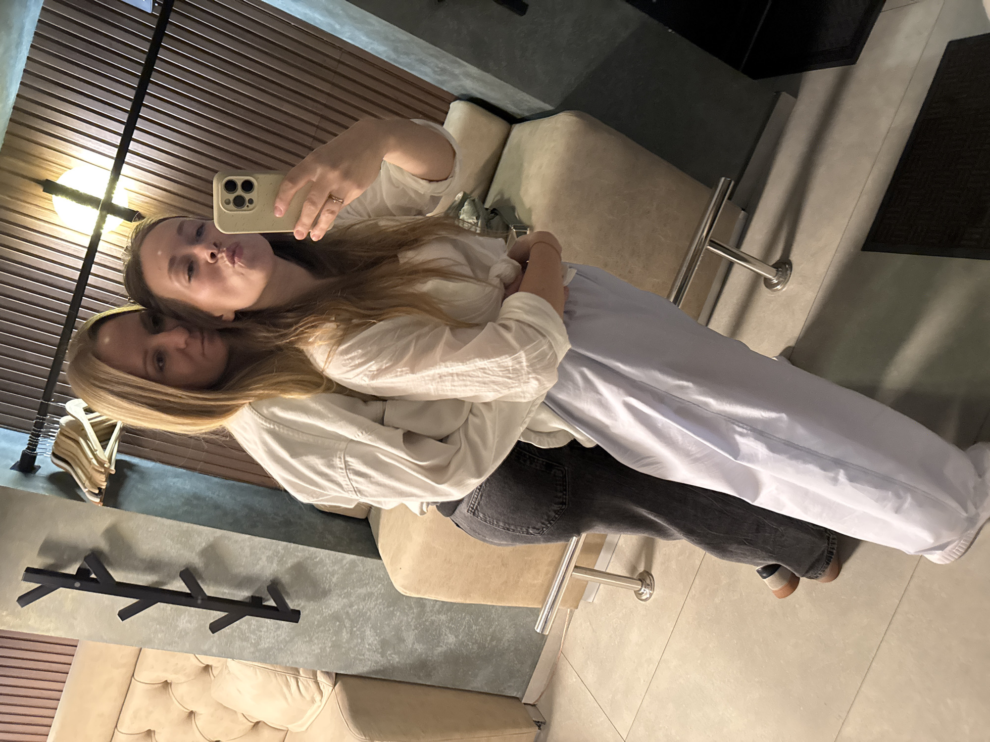 Two fair-skinned women are indoors, posing close together for a selfie. The woman holding the phone has brown hair and puckered lips. The other woman has long blonde hair. They both wear white shirts. The woman in front wears light blue sweatpants, while the woman behind wears dark grey jeans and brown shoes. Behind them is an interior setting featuring a light brown couch, a black coat rack, a coat rack attached to the wall, wooden-slatted walls, and light grey floors. The scene suggests a modern apartment or hotel room setting. While the city cannot be confirmed, the image could be from Budva, Montenegro.