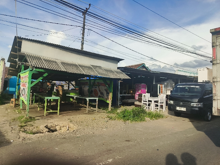 The scene is a street market in Cikalong, Indonesia (latitude: -6.74, longitude: 107.44). A large, open-sided shelter with a corrugated metal roof sits on the left side of the image, containing a food stall and two men. The stall is selling prepared food and drinks, with several tables and chairs arranged for customers. In the background, several buildings are visible, including a white building with a brown corrugated roof. A black van is parked on the right side of the image, facing away from the camera. The van has a white sticker on the back window. Several other vehicles are also parked in the background. There are many power lines running overhead throughout the scene. The ground is dirt and gravel, with a small ditch running through the middle of the image. There are some small bushes and trees growing in the foreground and middle ground. The sky is bright blue with some white clouds. The sun is shining and the weather is clear. The time of day is likely midday. The scene is typical of a busy street market in a developing country.