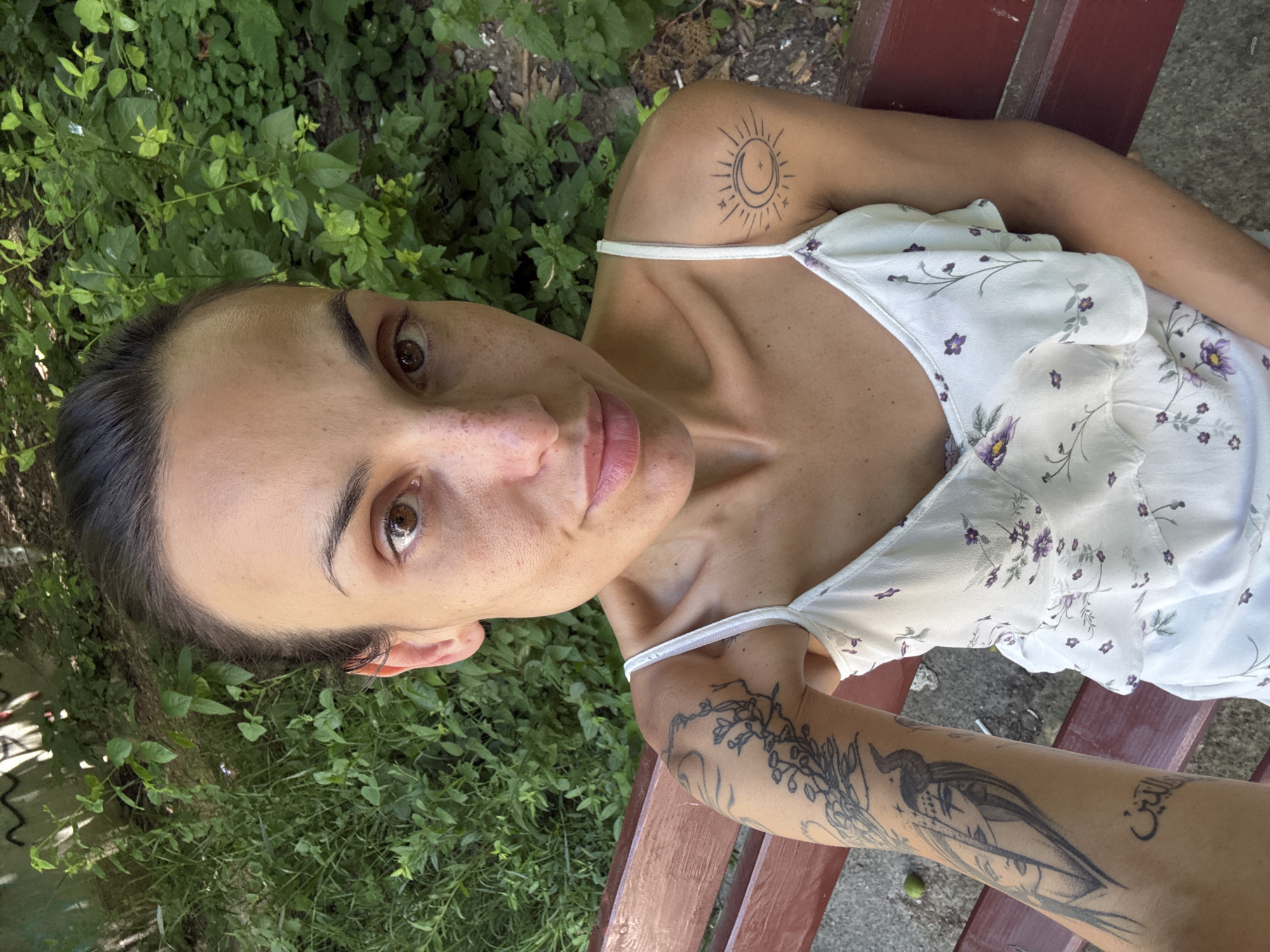 A selfie of a woman with dark hair pulled back, sitting on a wooden bench outdoors. She is wearing a white, floral-patterned dress with thin straps. She has tattoos on her left arm, including a detailed illustration of a face with horns and Arabic script, and a sun and moon tattoo on her right shoulder. The background consists of lush green foliage and a concrete surface. The image captures a bright, sunny day.