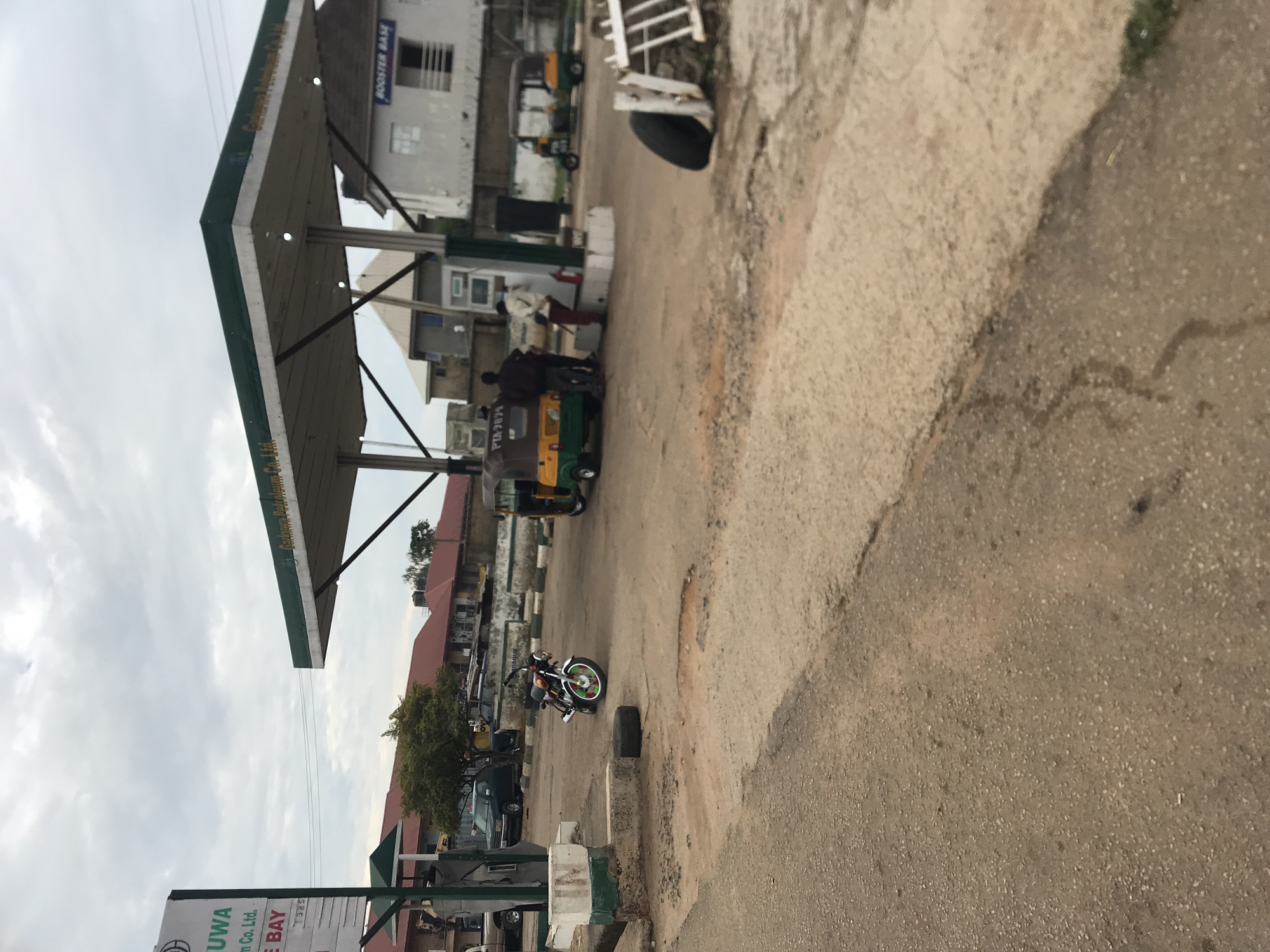 The image depicts an active gas station in Rafin Par, Nigeria, under an overcast sky. The central feature is a green and white canopy structure identifying "Garkuwa Petroleum Co. Ltd." Multiple fuel pumps are positioned beneath the canopy.
At the foreground and mid-ground, the ground consists of worn asphalt and dirt, showing various patches, cracks, and potholes. On the left, a large sign partially visible reads "GAR...UWA Petroleum Co. Ltd." with "BAY" and a partial number "7385" below.
Several vehicles are present. A yellow and green motorized tricycle, commonly known as a Keke Napep, with license plate "PIA 7079," is positioned at a fuel pump. Two individuals stand near this tricycle; one appears to be a pump attendant wearing a light-colored top and red trousers, facing the pump. Another person in dark clothing stands beside the tricycle. Further in the background, to the right, another yellow and green tricycle is visible. A motorcycle, predominantly dark with green and red accents on its front wheel spokes, is parked on the ground to the left of the pumps, near a loose tire lying flat. A dark-colored sedan is parked behind the station pumps, and a light-colored pickup truck is partially visible on the far left.
In the background, various buildings are visible, including structures with reddish-brown roofs and a prominent white building to the right with a blue sign that reads "BOOSTER BASE." A green tree stands in the mid-ground. Overheard power lines are faintly visible against the grey sky. On the far right, a tire leans against a white, possibly broken, fence or structure.