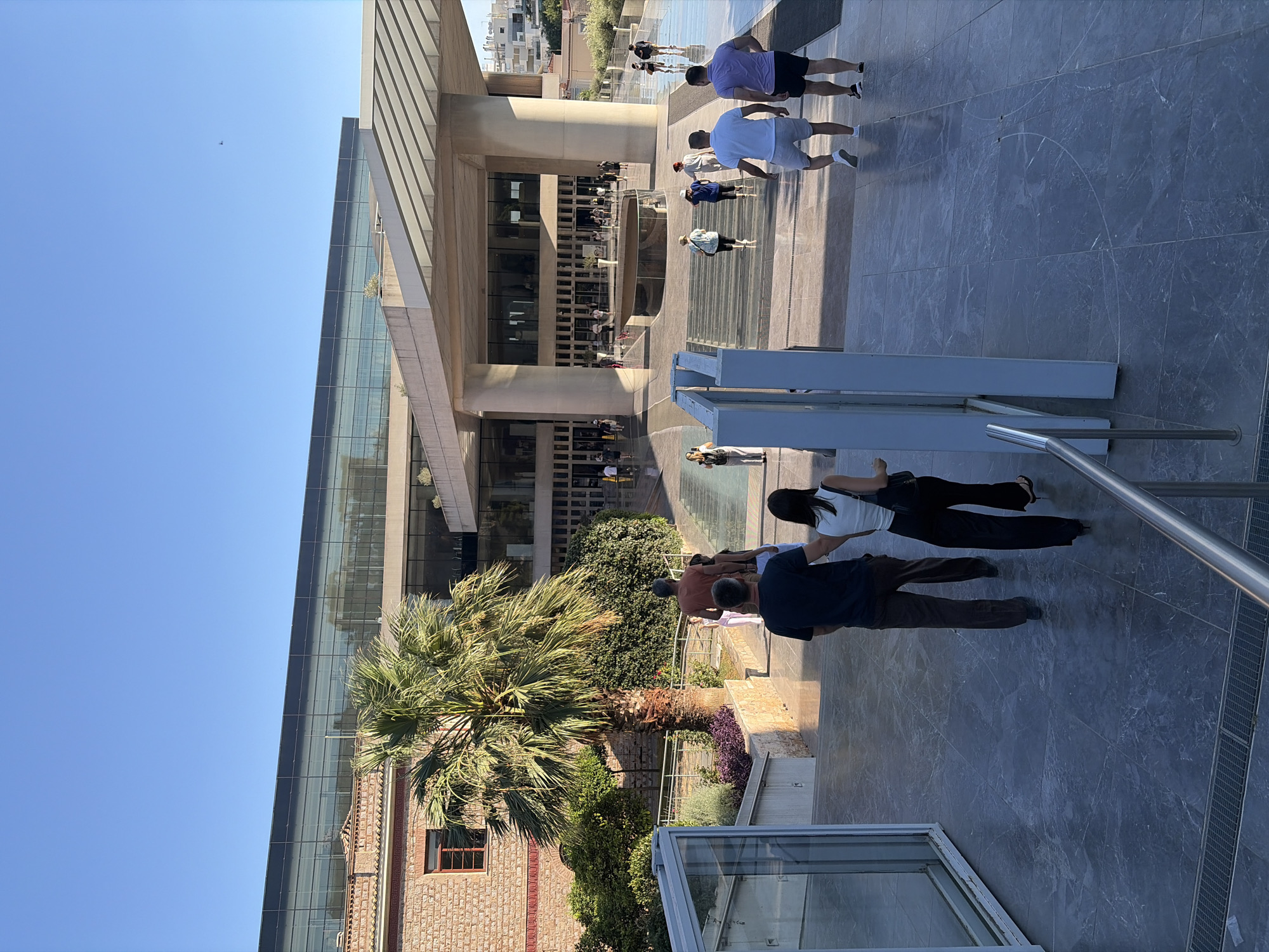 The image shows a group of people walking along a paved walkway in front of a modern building with large glass windows. The building's architecture is contemporary, with clean lines and geometric shapes. Some people are dressed casually, suggesting a warm day, possibly afternoon, given the bright sunlight casting shadows. The background reveals a clear blue sky, indicating good weather.
The scene appears to be a public space, possibly a museum or a cultural center, given the architectural style and the presence of visitors. Some individuals are walking towards the camera, while others are in the distance, interacting with the environment. A palm tree is visible in the mid-ground, adding a touch of greenery and suggesting a Mediterranean climate, consistent with the provided location context of Athens, Greece.
Notable details include the varied attire of the people, ranging from t-shirts and shorts to more formal wear. The ground is covered in large, grey tiles with a marble-like pattern. A metal railing is visible on the right side of the image, and a section of a metal detector frame is present in the foreground, hinting at security measures in place. There is no visible text in the image. The overall mood seems relaxed and leisurely, with people enjoying their time outdoors.