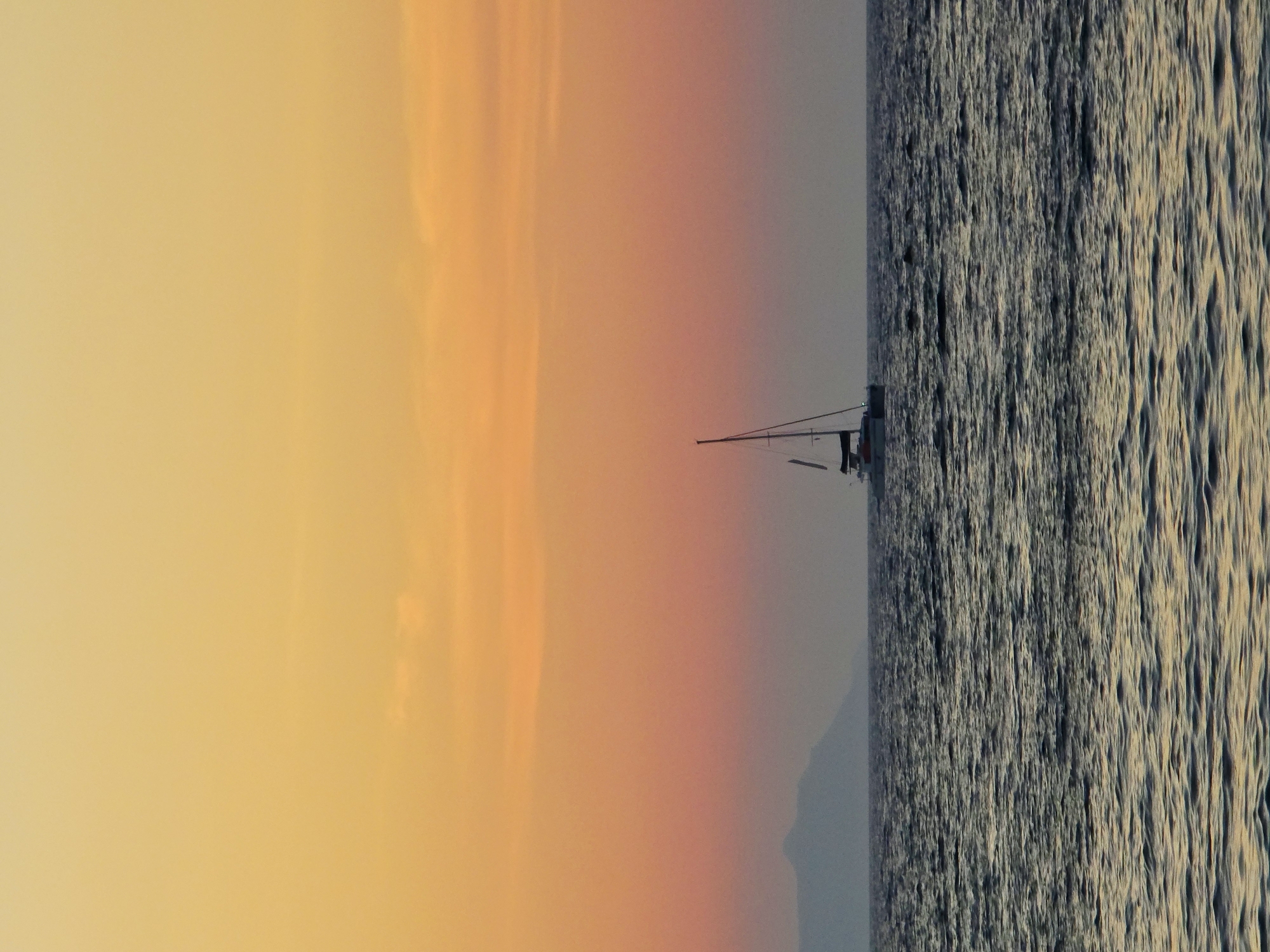 The image depicts a seascape with a catamaran sailing on the water during sunset. The sky is a gradient of warm colors, transitioning from yellow at the top to orange and pink closer to the horizon. In the distance, a faint silhouette of mountains can be seen. The water is choppy with visible waves. The catamaran is the only prominent object in the water, with its mast and sails visible. The scene evokes a sense of calm and tranquility. It is not possible to determine the specific location within Chania, Greece, from the image alone.