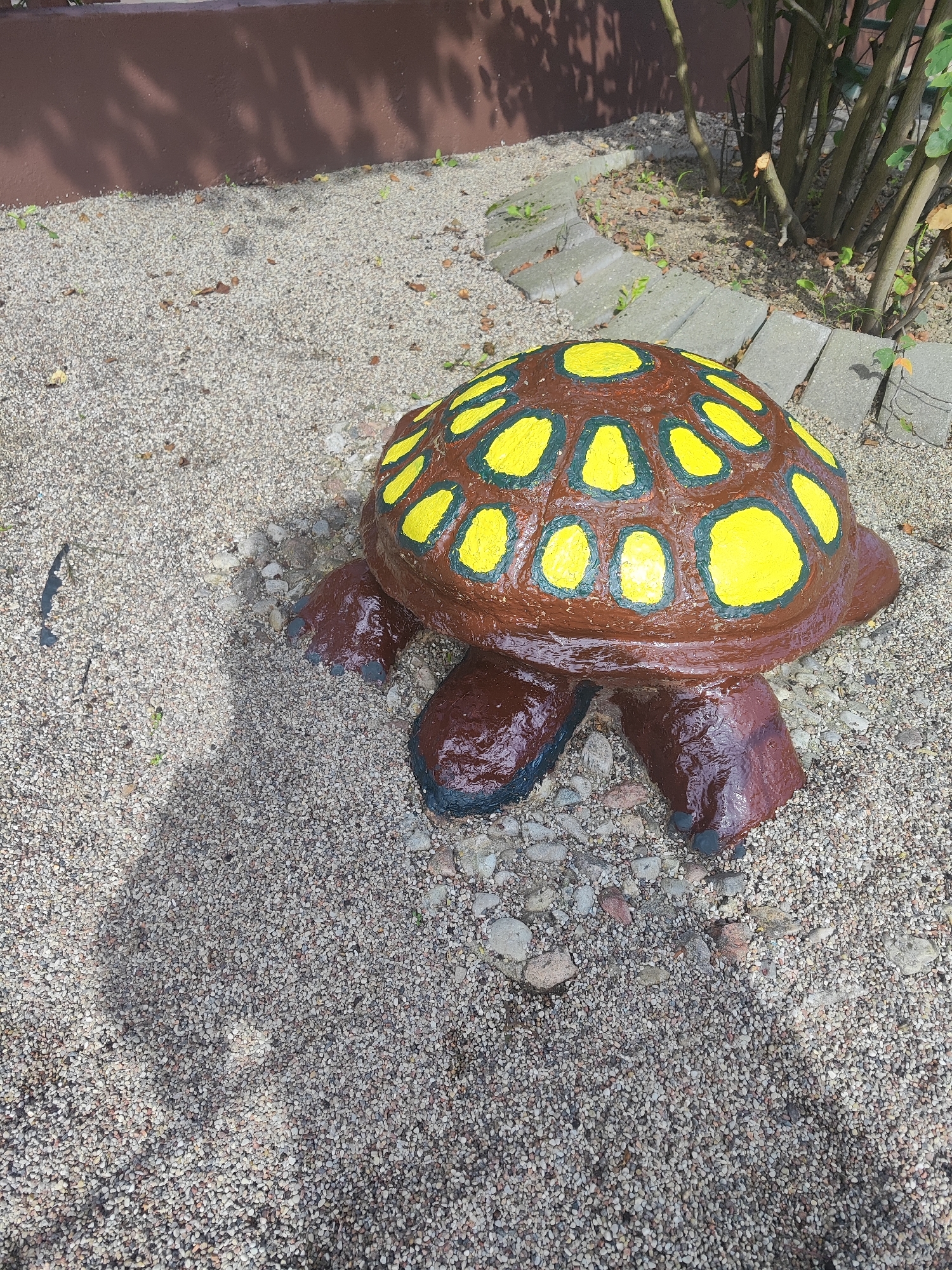A concrete garden decoration shaped like a turtle is placed on a gravelly ground. The turtle's shell is brown with yellow oval markings outlined in dark green. Its legs and head are a dark reddish-brown, with dark gray claws visible on the front legs. A shadow of what appears to be tree branches is cast across the ground from the top left to the bottom center of the image. The background features a brown wall with more tree branches and a pathway made of rectangular concrete pavers curving around the base of a bush. The environment appears to be outdoors, likely a garden or yard.