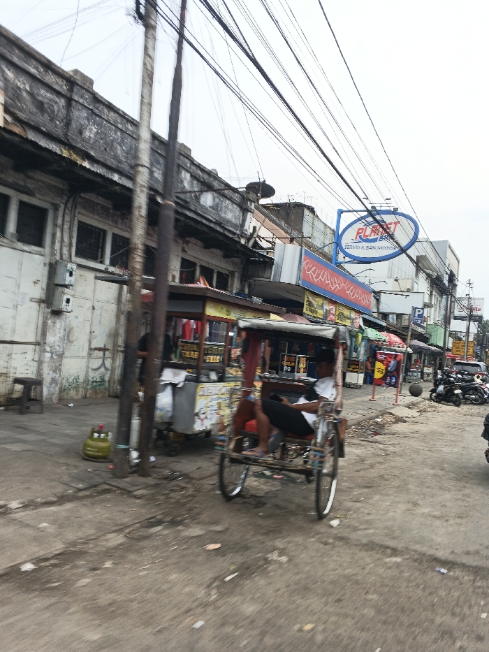 The image depicts a daytime street scene in Bandung City, Indonesia, characterized by urban infrastructure and commercial activity under an overcast sky. In the foreground, a paved but dirty street runs diagonally from bottom left to top right, showing debris and discolored patches. A traditional three-wheeled pedicab (becak) occupies the mid-ground, traveling right to left. A male operator, wearing a white short-sleeved shirt and dark pants, is seated in the driver's position, facing slightly left.
To the left of the pedicab, a portable vendor's stall with a canopy and displayed merchandise (text not fully legible) is positioned on the sidewalk, partially obscured by a dark utility pole. Another utility pole stands prominently in the center foreground. A green LPG tank and a small wooden stool are visible on the sidewalk to the far left.
The left side of the image features a weathered, multi-story building with dark, closed windows and a dilapidated facade. Overhead, numerous black electrical or communication wires crisscross the sky, attached to utility poles.
In the mid-ground to the right, a bright blue oval sign with "PLANET SERVIS & BAN MOTOR" in white text is mounted above a row of commercial establishments. These buildings display various signs in red, yellow, and blue, indicating shops or services. Further down the street, on the right, several motorcycles are parked in a cluster. The street extends into the background, where more buildings and parked vehicles are visible, fading into the distance.