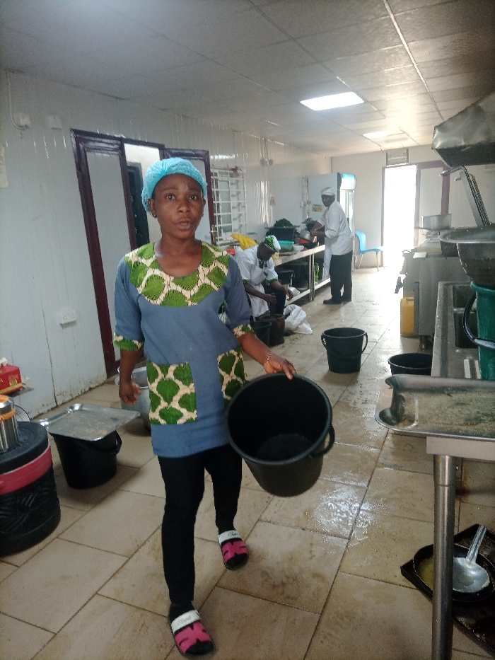 In a commercial kitchen, a woman in a blue and green patterned tunic, black leggings, and sandals carries a black bucket. She wears a blue hairnet and looks towards the camera. Behind her, other workers in chef's whites are preparing food at a counter. The room is brightly lit with tiled floors and stainless steel equipment. Black buckets are scattered throughout the space. A doorway leads to another room filled with light.