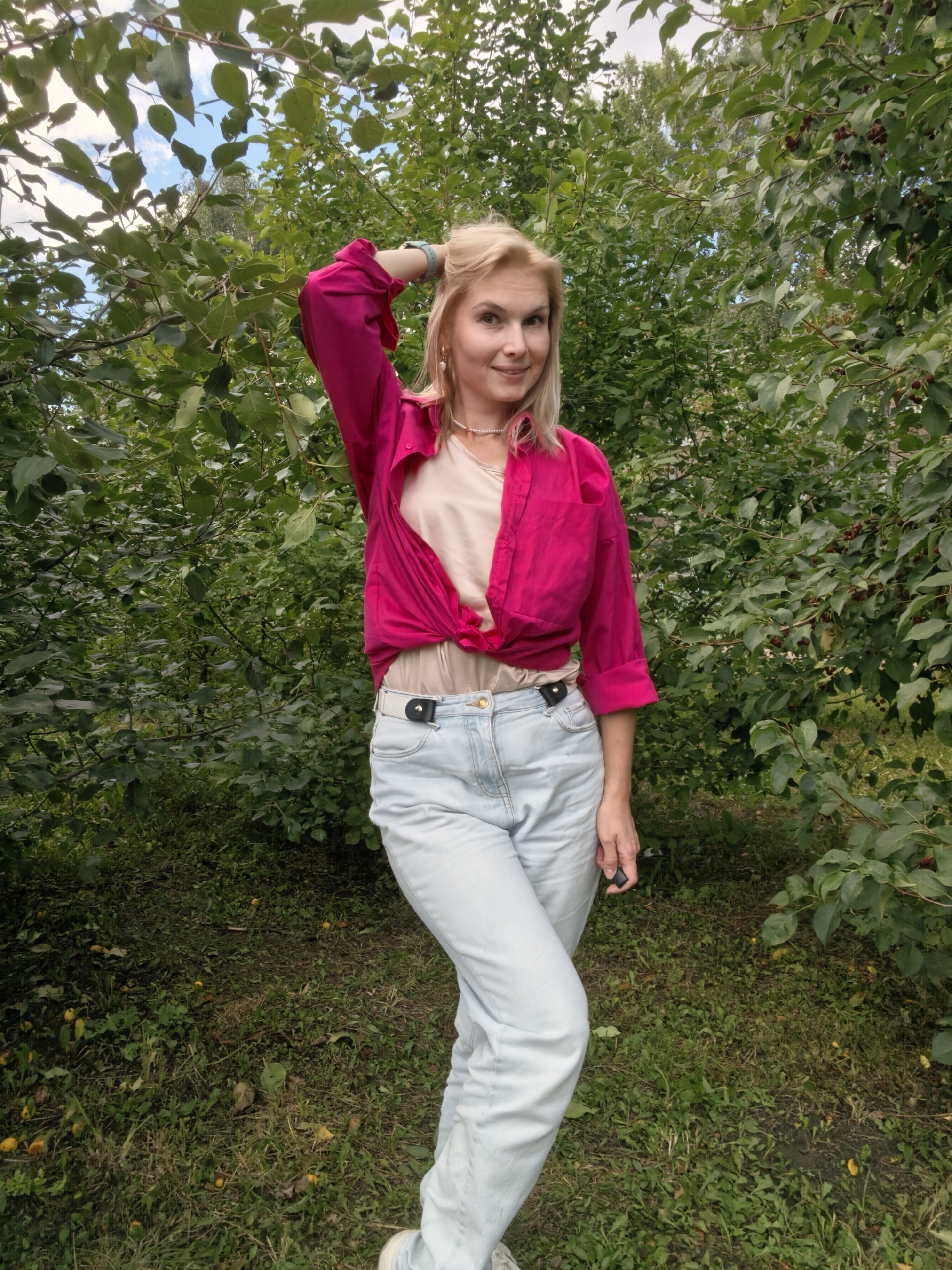 A blonde woman in a pink shirt and light blue jeans stands outdoors in a leafy environment. She has her left hand on her head and is looking at the camera with a slight smile. Her shirt is open and tied at the waist, revealing a beige top underneath. She is wearing a white belt and white sneakers. The background is filled with lush green foliage and trees, suggesting a park or garden setting. The ground is covered with grass and some fallen yellow leaves. The presence of fallen leaves and the overall greenery suggests it may be autumn. It is not possible to determine the specific location within Sterlitamak, Russia from the image.